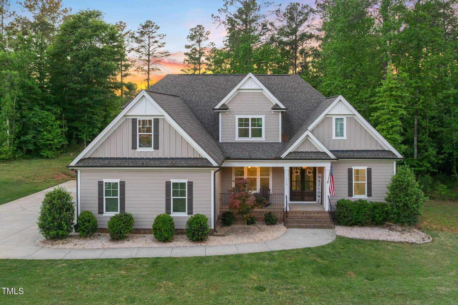 A two-story suburban house with beige siding, white trim, a covered front porch, dormer windows, and neatly landscaped shrubs sits at 175 Lockamy Lane in Youngsville, set against a background of trees at sunset. An American flag hangs near the entrance.