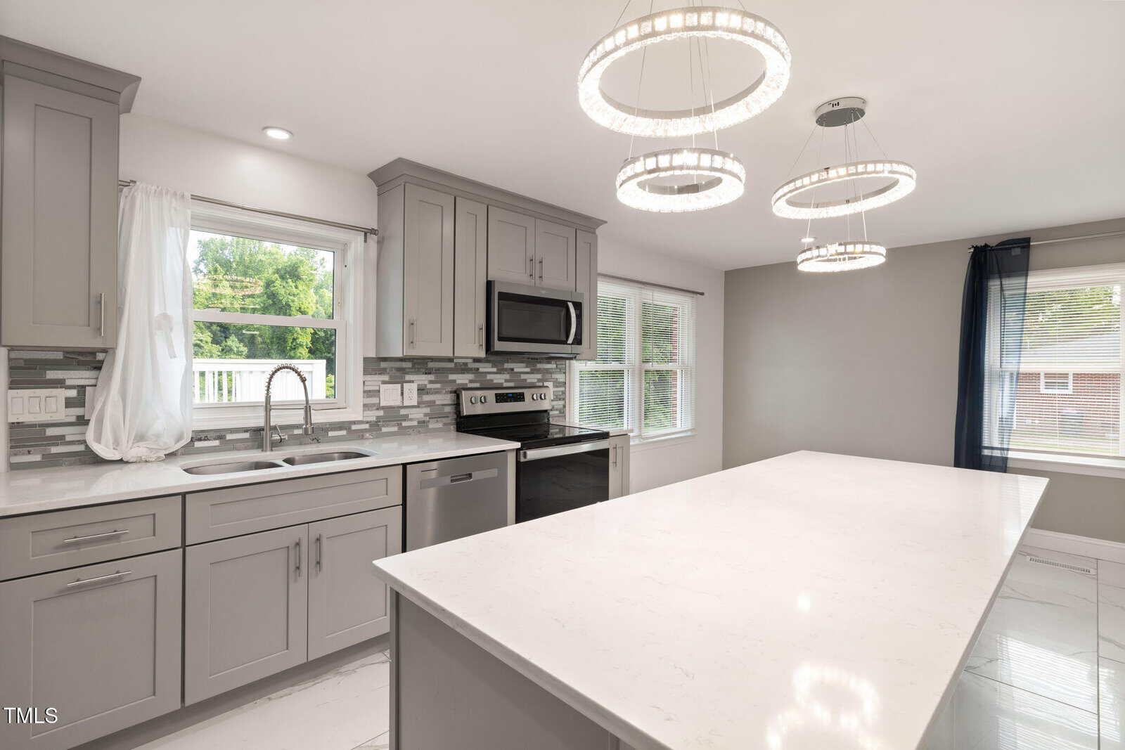 Modern kitchen at 2315 Oxford Road, Henderson features gray cabinets, stainless steel appliances, white marble countertops, a large island, tile backsplash, and three circular pendant lights. Large windows provide ample natural light.