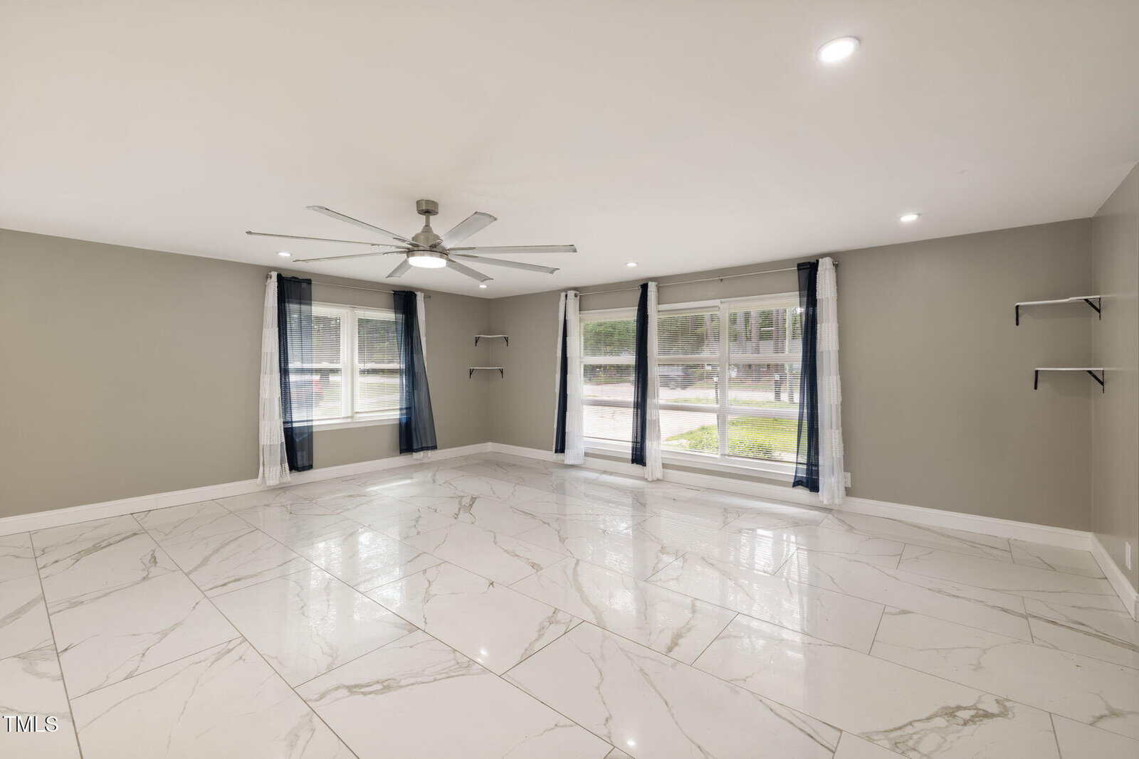 A spacious, empty room with large white marble floor tiles, gray walls, two large windows with dark curtains, a ceiling fan with lights, and wall-mounted shelves on either side.