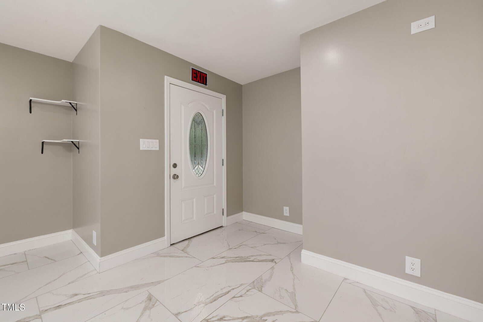 A small entryway with light gray walls, white crown molding, large white marble tile flooring, a white door with an oval window, two black wall shelves, and an exit sign above the door.