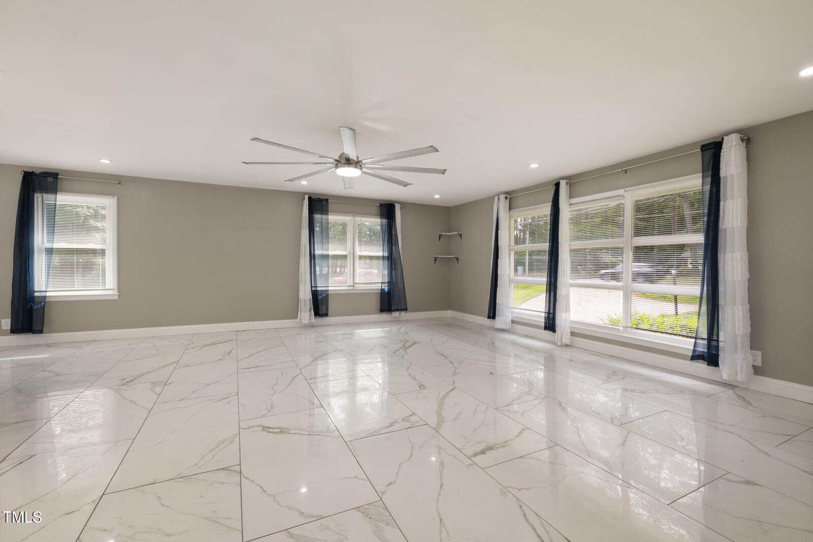 Spacious empty room with large white marble floor tiles, gray walls, a ceiling fan with metal blades, several windows with blue and white curtains, and recessed lighting in the ceiling.