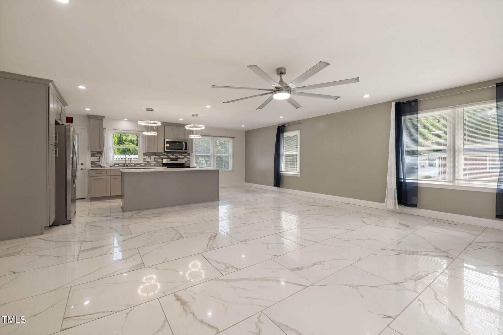 Spacious open-plan kitchen and living area with large white marble tile flooring, ceiling fan, recessed lighting, and windows with dark curtains allowing natural light into the room. Kitchen features modern appliances and an island.