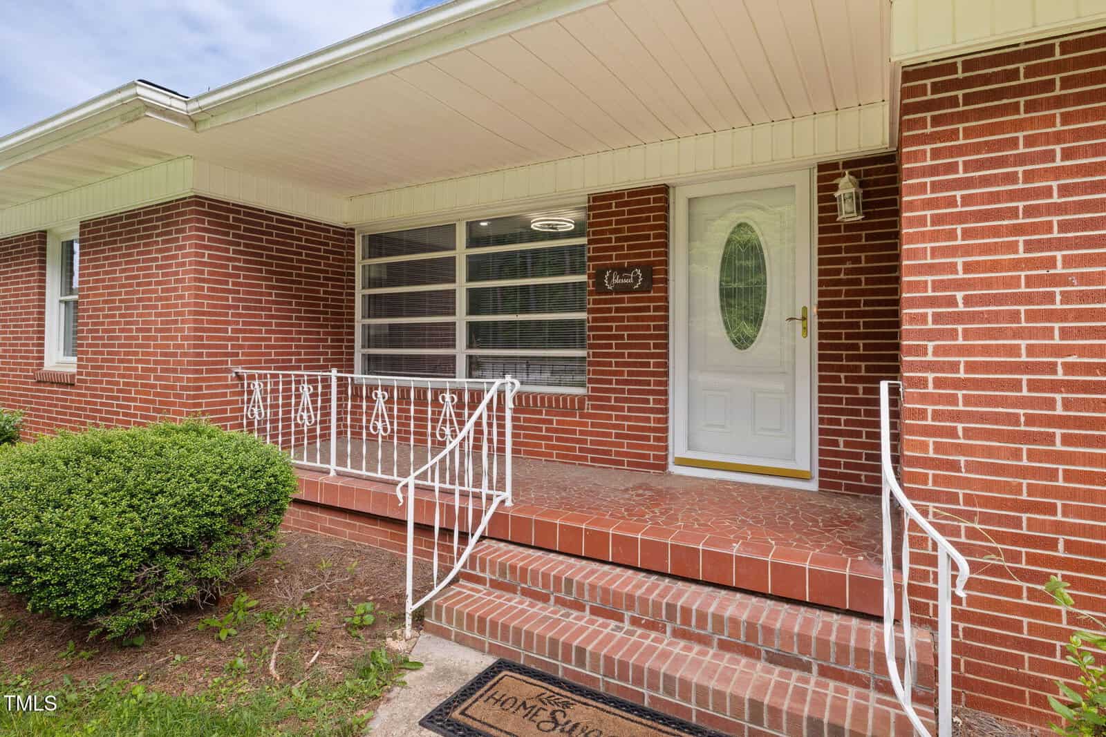 Front porch of a brick house with a white door featuring an oval window, white metal railing, red-tiled steps, and a doormat that says "Home Sweet Home." Bushes and grass are visible beside the steps.