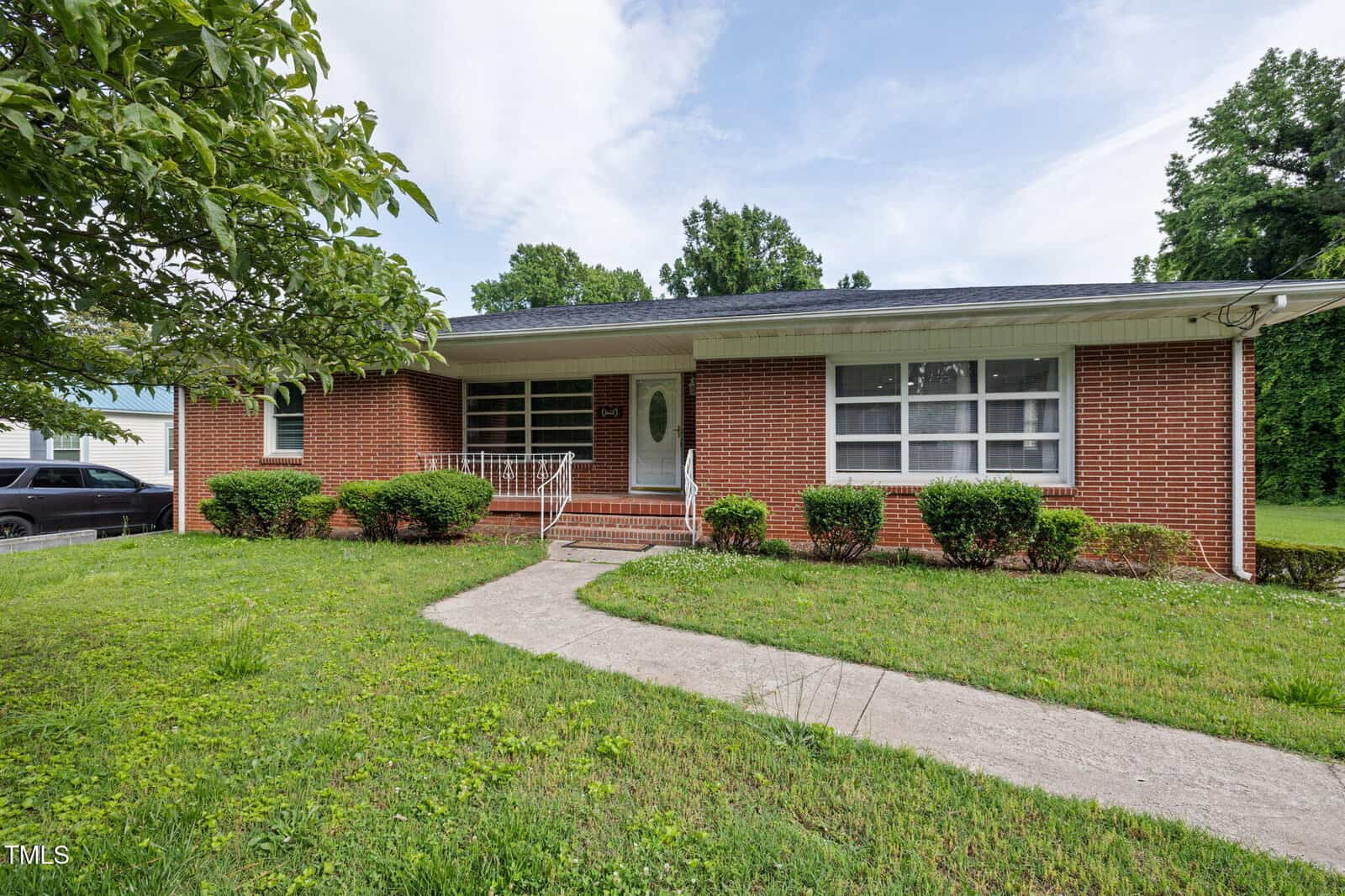 Single-story red brick house with a covered front porch, white trim, and a light-colored front door. A concrete walkway leads to the entrance, with neatly trimmed bushes and a grassy lawn in front.