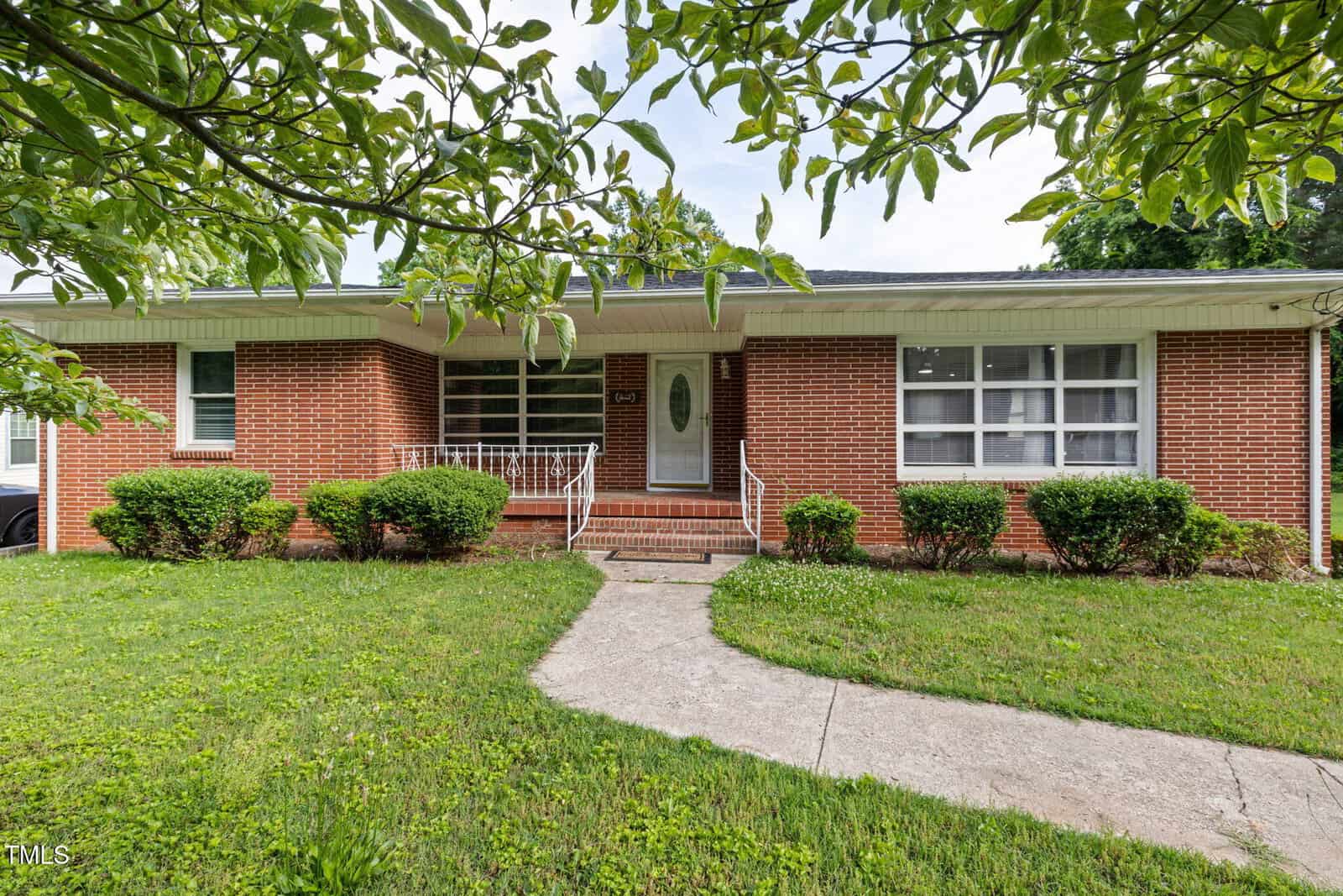 Single-story brick house with a small covered porch, white trim, and a white front door. Shrubs line the front of the house, and a concrete walkway leads to the entrance. Grass lawn and tree branches frame the scene.