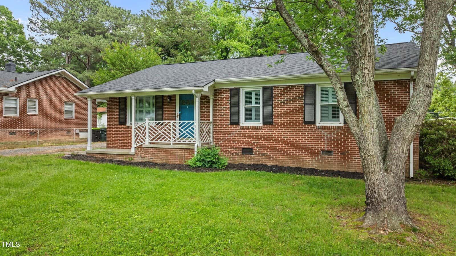 A one-story red brick house with black shutters, a covered front porch with white railing, and a blue front door. There is a green lawn, a tree in the yard, and another brick house partially visible on the left.