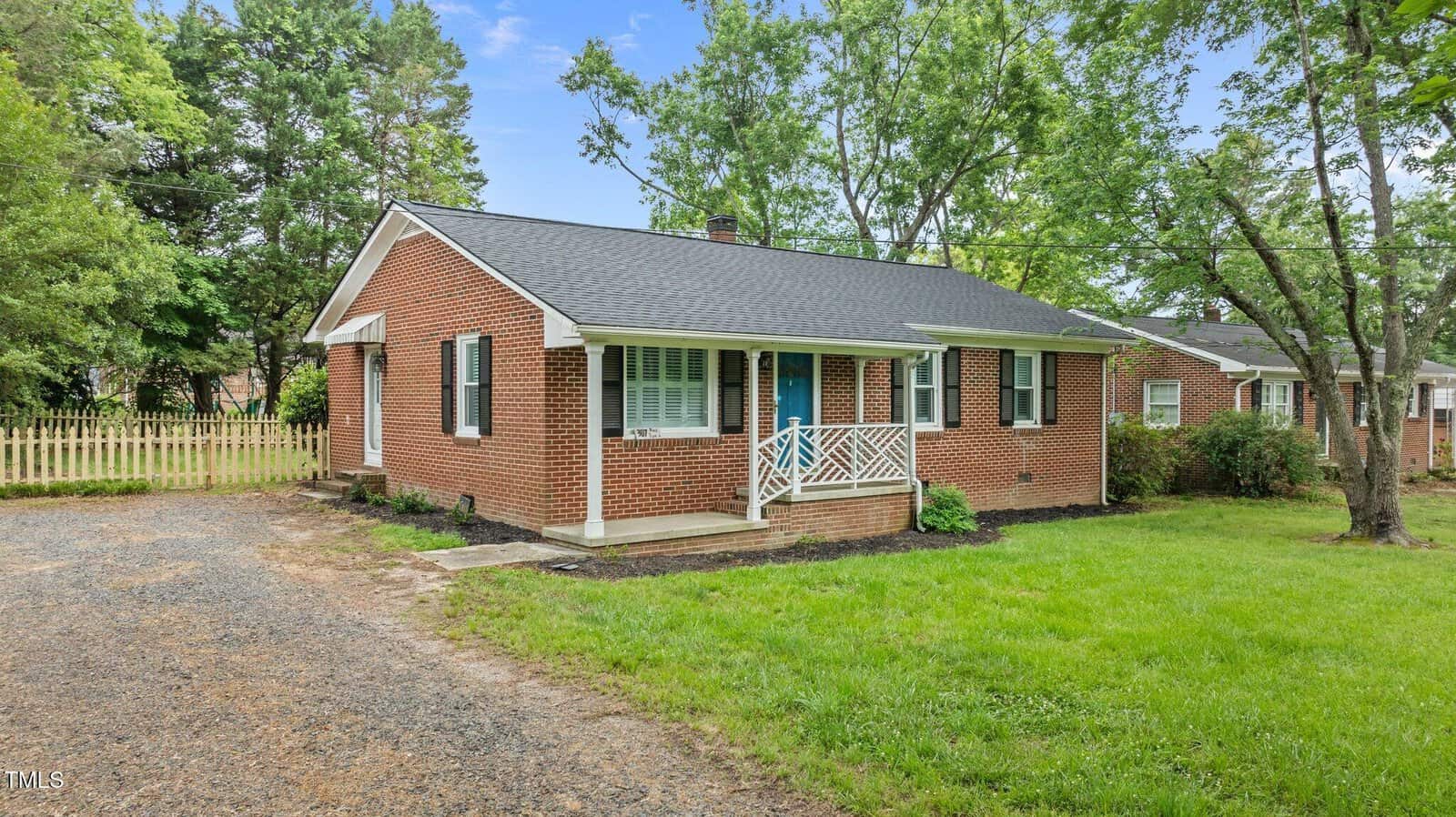 A single-story brick house with a dark shingled roof, blue front door, and white trim sits on a grassy lawn. A small porch with white railings is at the entrance. A gravel driveway runs along the side of the house.