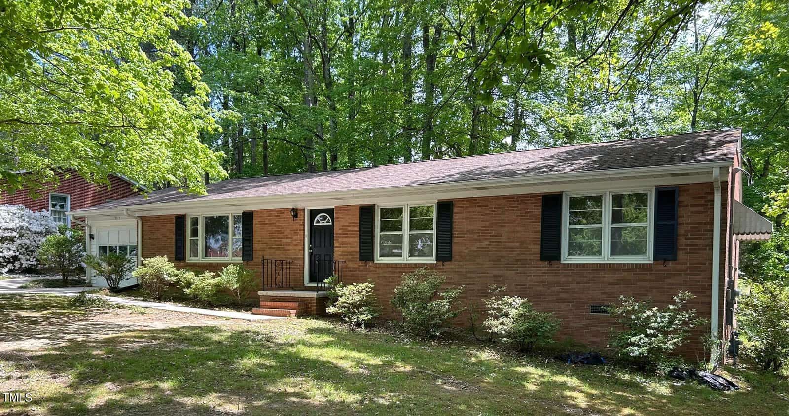A single-story brick house with white-framed windows and black shutters, surrounded by trees and shrubs. There are steps leading to a black front door, and a driveway is visible to the left.