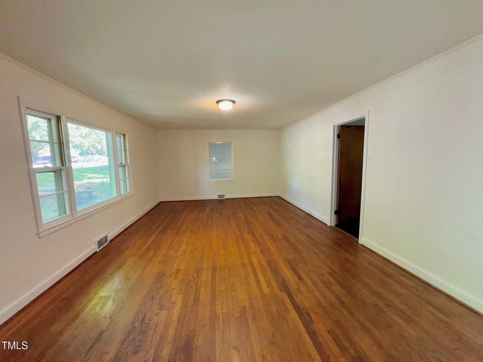An empty room with hardwood floors, white walls, a ceiling light, three windows on the left wall, a single window on the far wall, and a doorway on the right leading to another space.