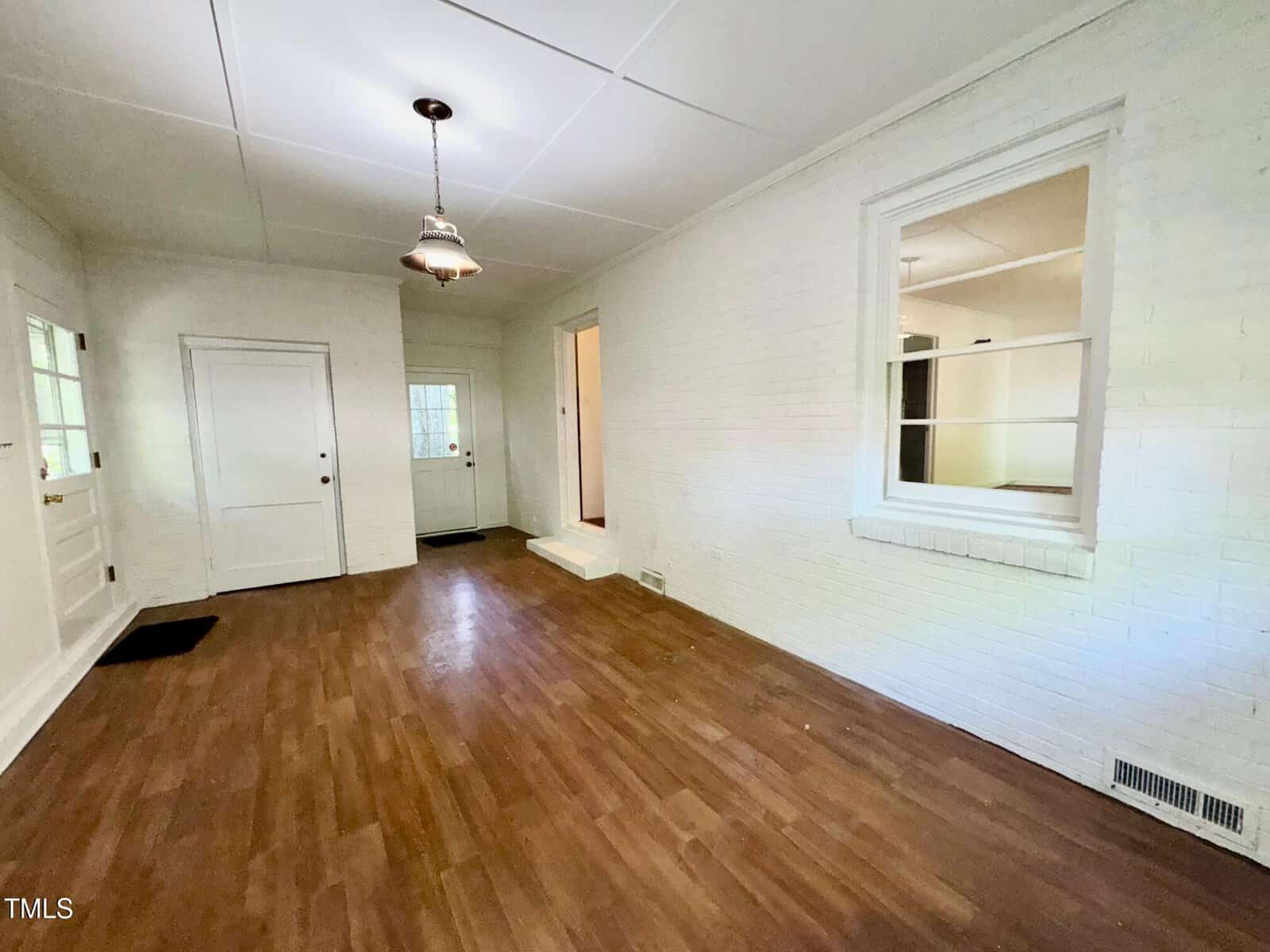 A rectangular room with white brick walls, wood flooring, a ceiling light fixture, two doors, a window opening to another room, and a small step leading to a hallway. The space is empty and well-lit.