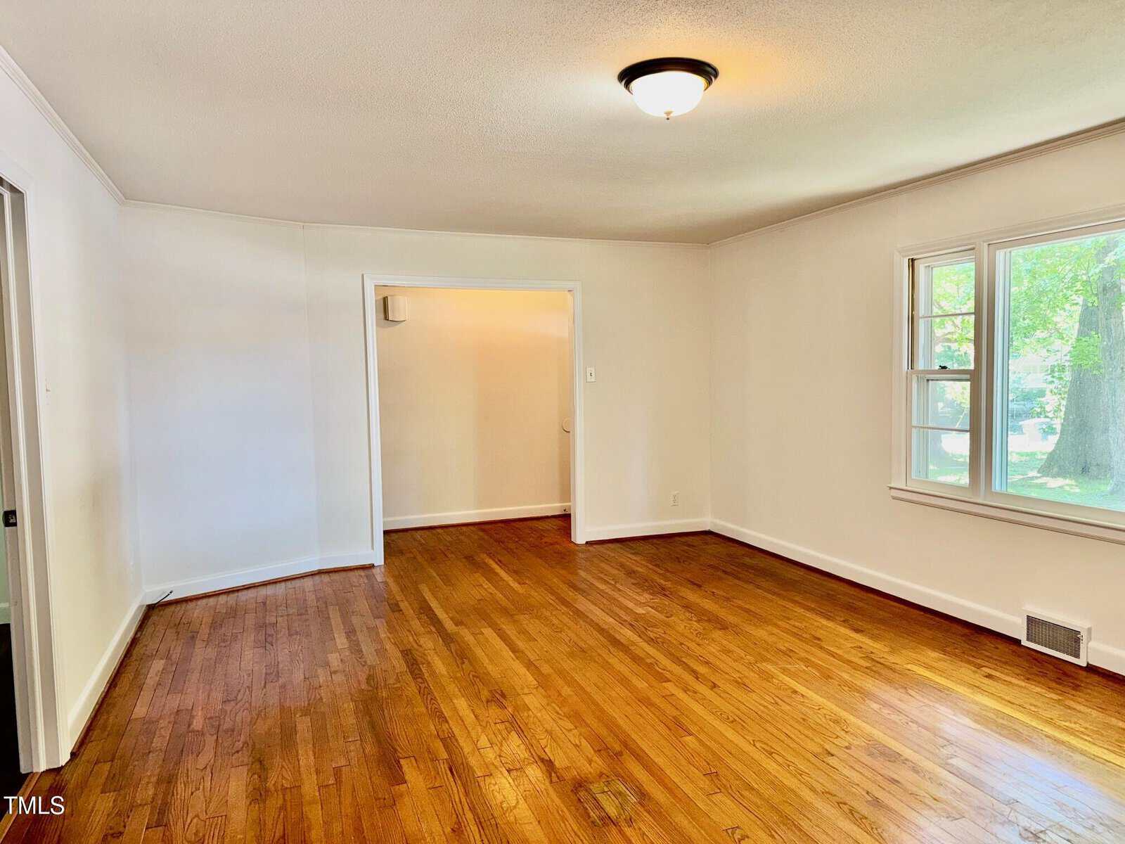 Empty room with hardwood floors, cream-colored walls, a ceiling light, a window letting in natural light, and an open doorway leading to another space. A floor vent is visible near the window.