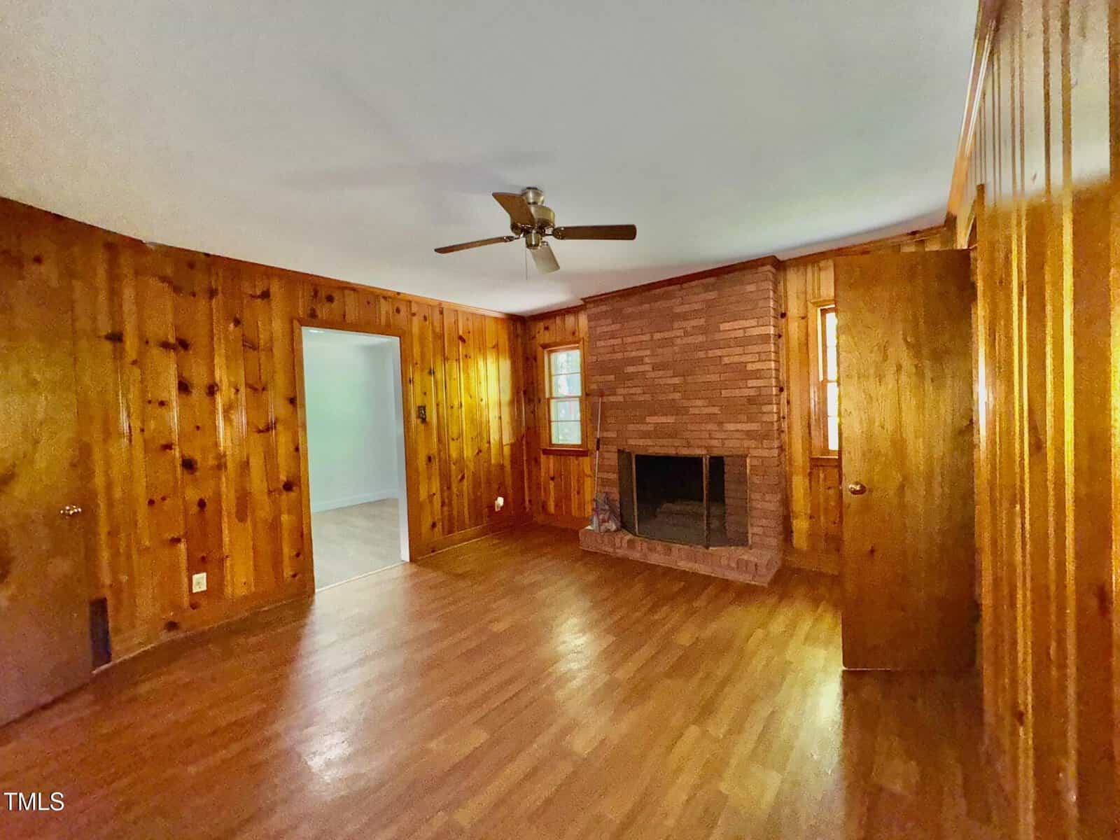 A wood-paneled living room with hardwood floors, a ceiling fan, a brick fireplace, two small windows, and two closed wooden doors. An open doorway leads to an adjacent room.