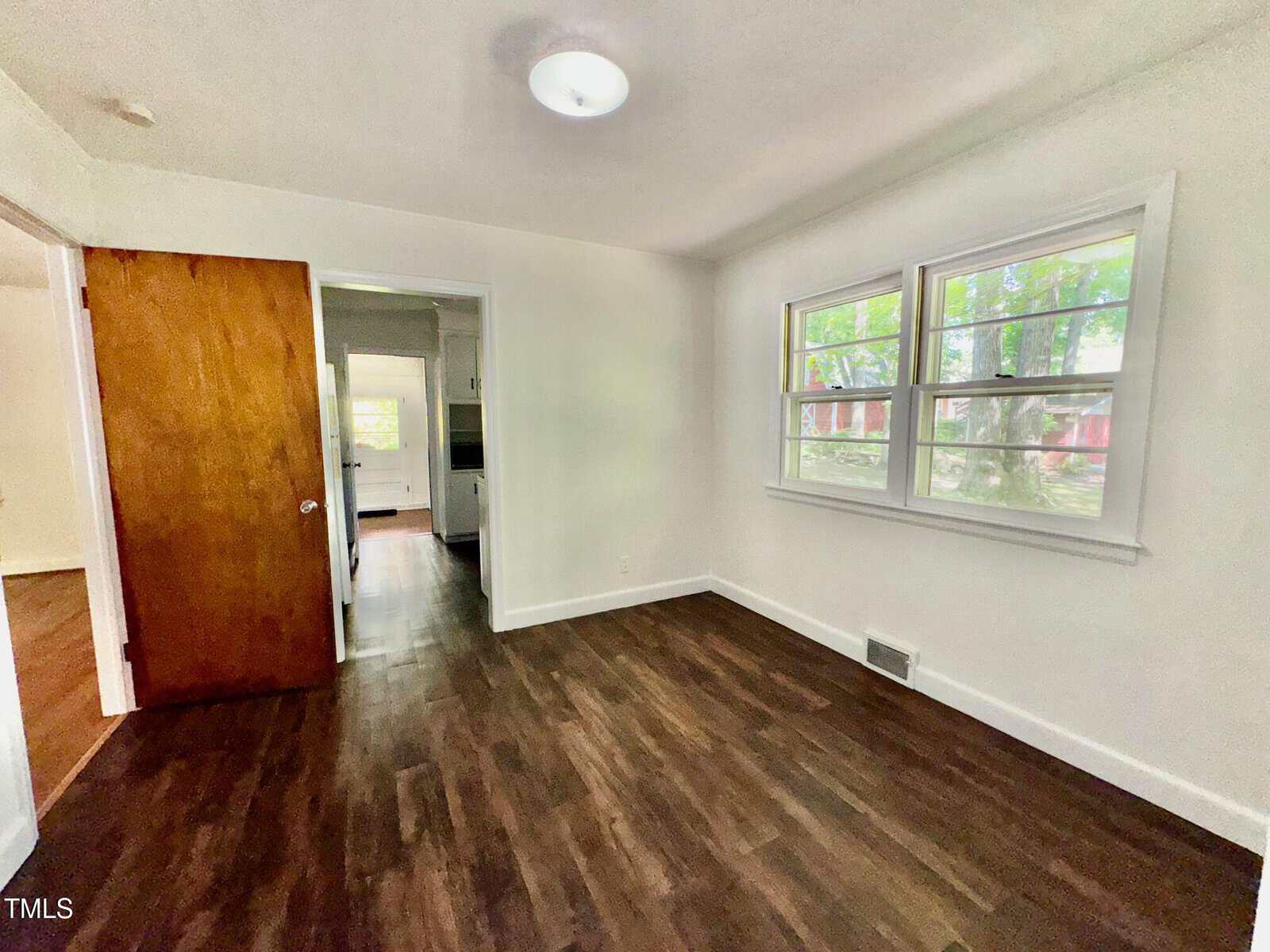 A small, empty room with white walls, dark wood flooring, a wooden door, and two windows letting in natural light. The room connects to a hallway and another space visible through the door.