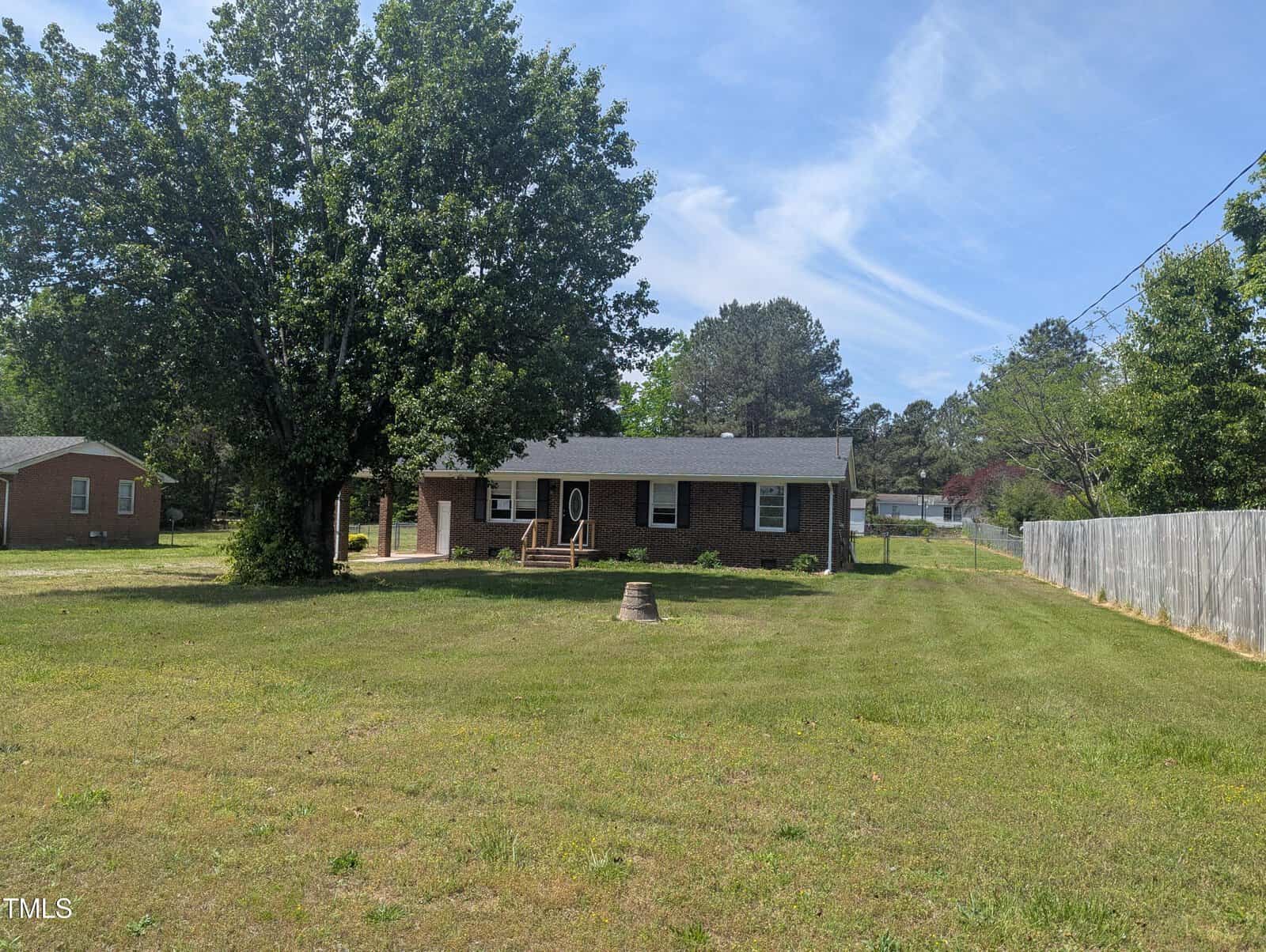 A one-story brick house with a covered front porch sits on a large grassy lot. A tree stands to the left of the house, and there is a stump or structure in the front yard. A wooden fence lines the right side.