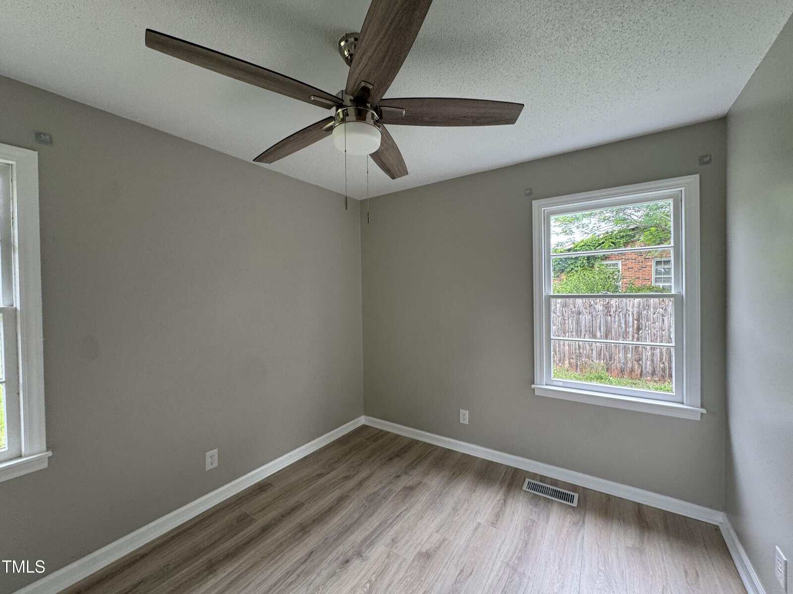 Small empty room with two windows, light gray walls, wood-patterned floor, a ceiling fan with wooden blades, and a view of a brick building and wooden fence outside.