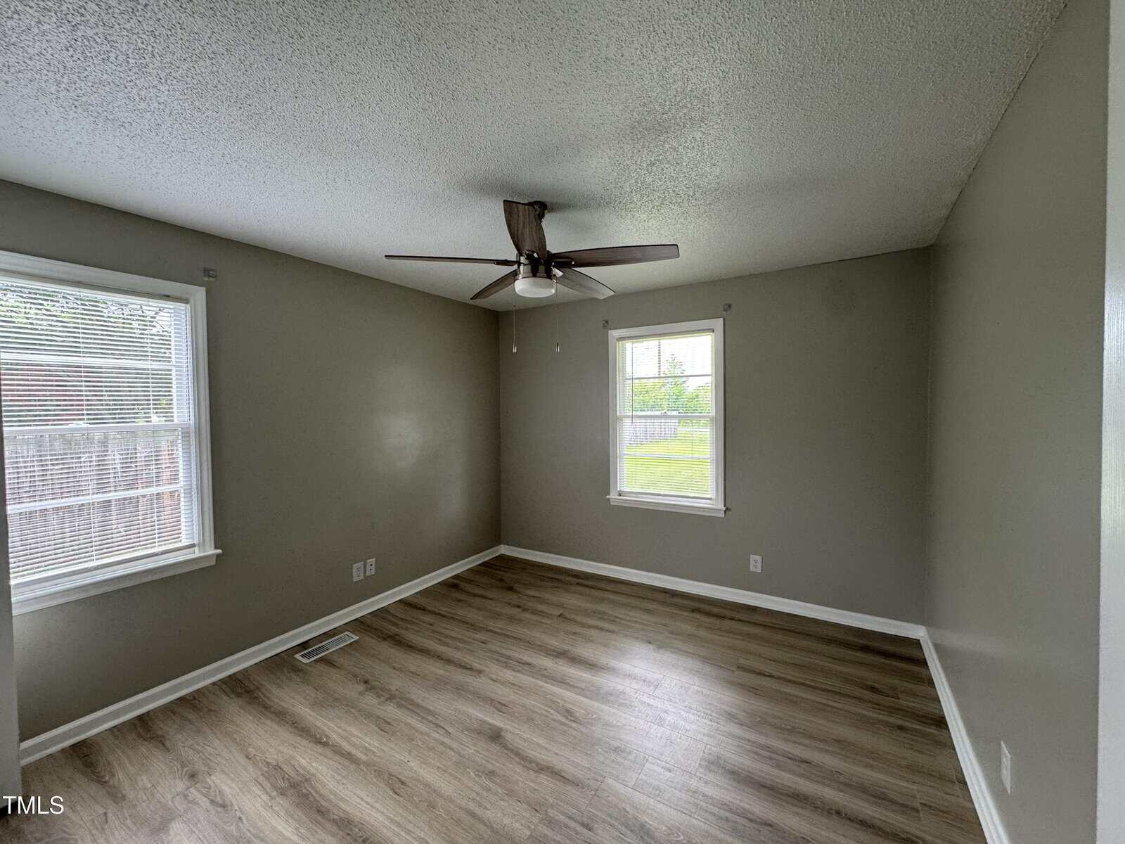 Empty room with light gray walls, light wood flooring, two windows with blinds, a ceiling fan with light, and a white baseboard along the walls. Natural light enters through the windows.