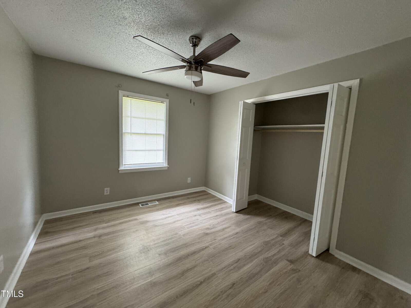 Empty room with light wood flooring, beige walls, a ceiling fan, a window with closed blinds, and an open closet with white doors. Natural light is coming in through the window.