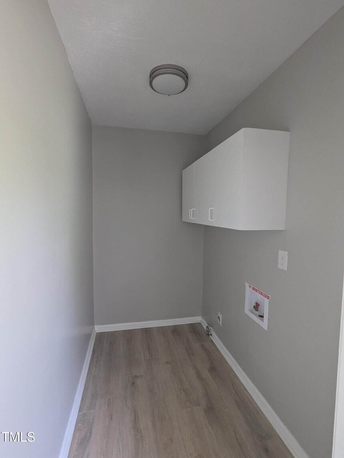 A small, narrow room with light gray walls, light wood flooring, a flush-mounted ceiling light, a white wall cabinet, and utility connections for laundry on the right wall.