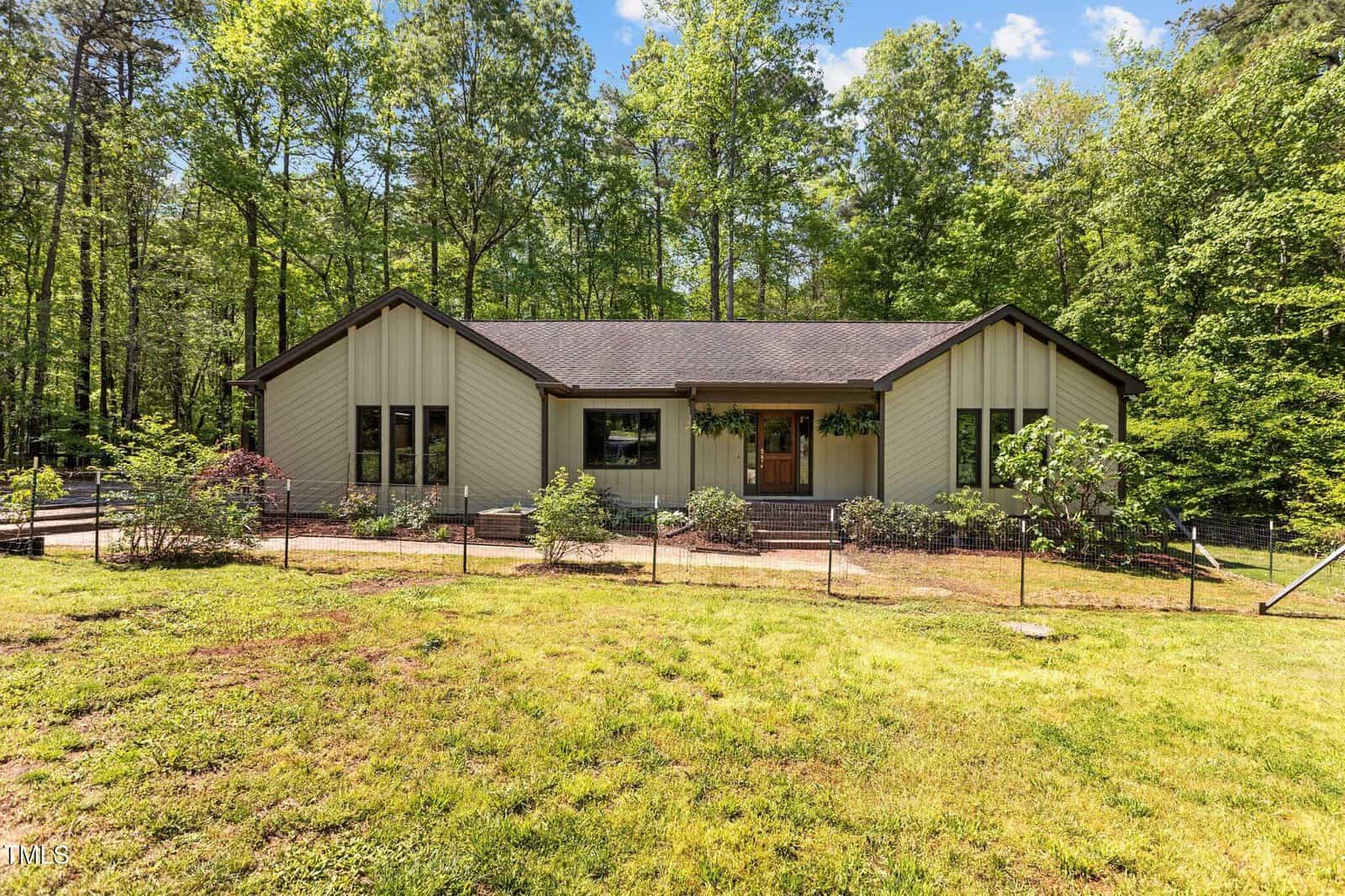 A single-story beige house with dark trim sits in front of a wooded background. The home has large windows, a fenced yard, and steps leading to the front entrance, surrounded by grass and greenery.