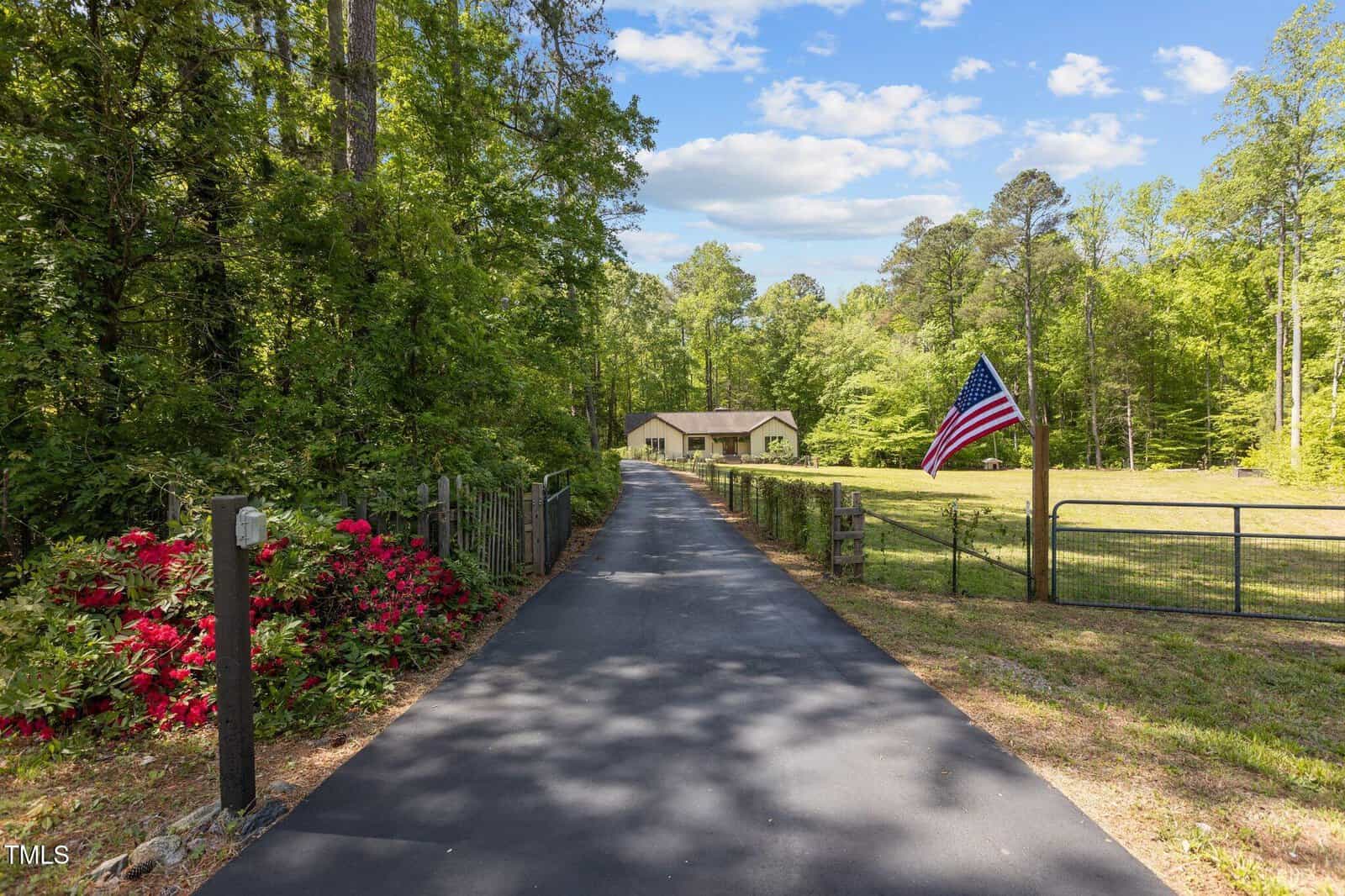 A paved driveway leads to a house surrounded by trees and greenery, with an American flag on a pole by a fence. The sky is partly cloudy and colorful flowers bloom by the driveway.