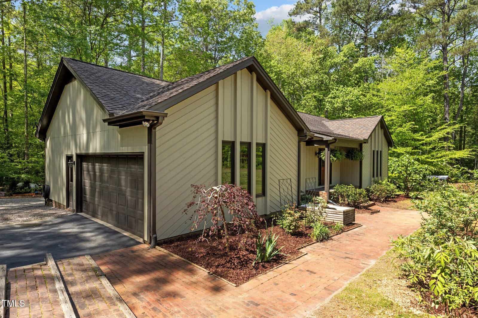 Modern single-story house with light-colored vertical siding, dark trim, and a two-car garage, surrounded by lush trees. A brick walkway leads to the front entrance with landscaped plants along the path.