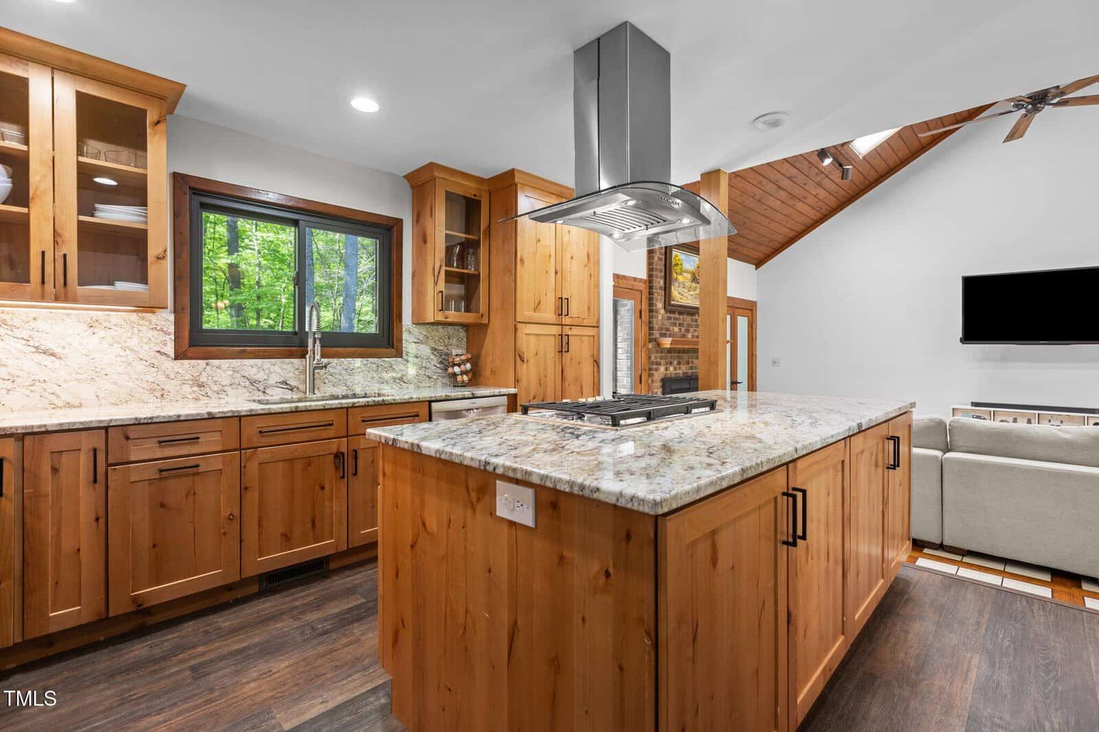 A modern kitchen with wooden cabinets, a marble countertop island featuring a built-in gas cooktop, stainless steel range hood, dark wood floors, and a window showing green trees. An open living area is partially visible.