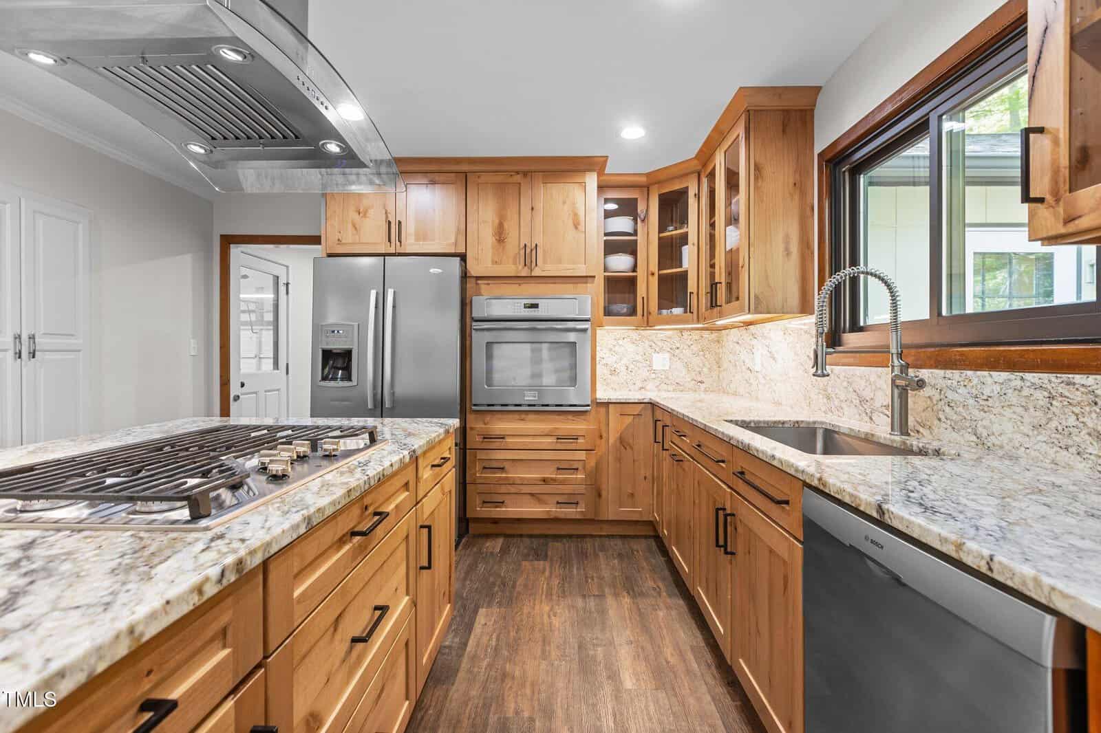 Modern kitchen with wood cabinets, granite countertops, a stainless steel refrigerator, built-in oven, stovetop island, dishwasher, deep sink, and large window. Dark wood floors and under-cabinet lighting are visible.