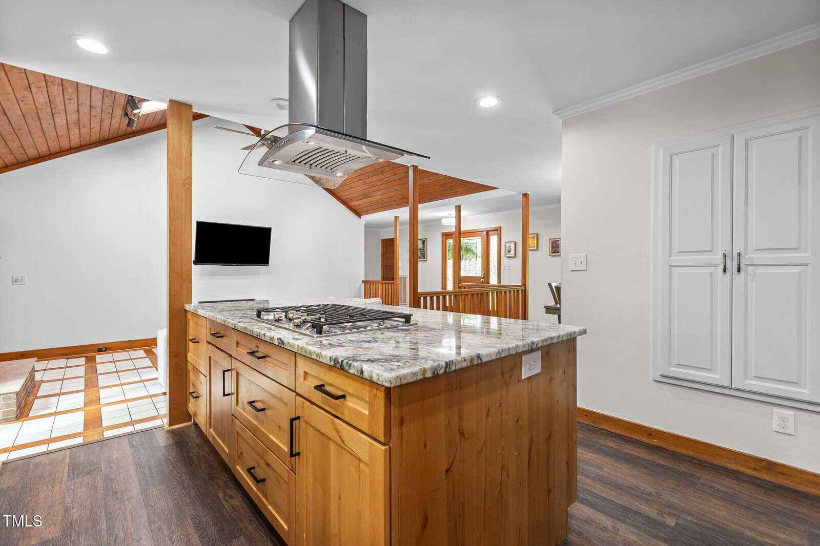 Modern kitchen with a wood island featuring a built-in gas cooktop and stainless steel hood. The space has wood flooring, white walls, ceiling lights, and a small white cabinet on the right wall.