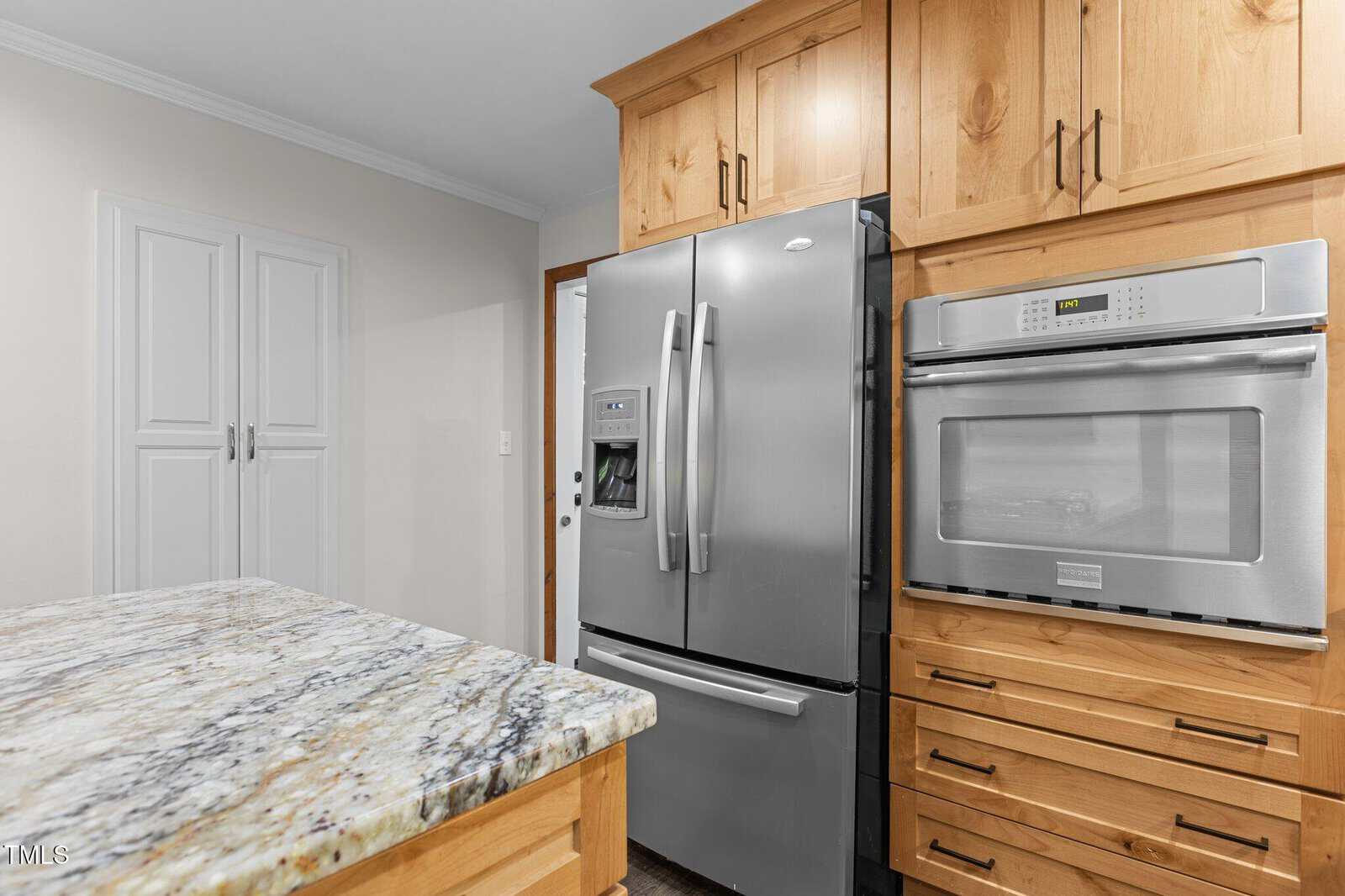 A stainless steel French door refrigerator and a built-in oven are set in wooden cabinetry next to a granite kitchen island. White pantry doors are visible in the background.
