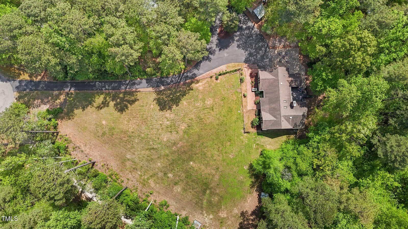 Aerial view of a house surrounded by dense trees, with a large, open grassy yard and a curved driveway leading to the home. The property is bordered by forested areas on multiple sides.
