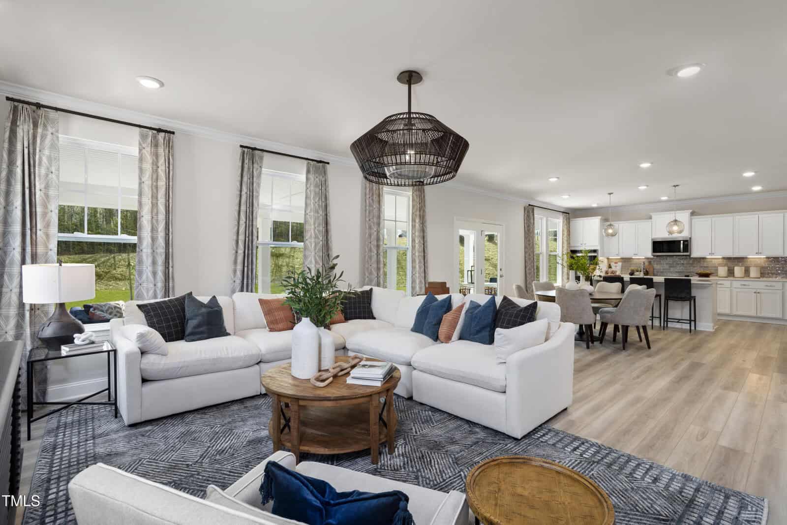 A spacious, modern living room with white sofas, decorative pillows, a round wooden coffee table, and large windows. The room flows into an open dining area and kitchen with white cabinets and pendant lighting.