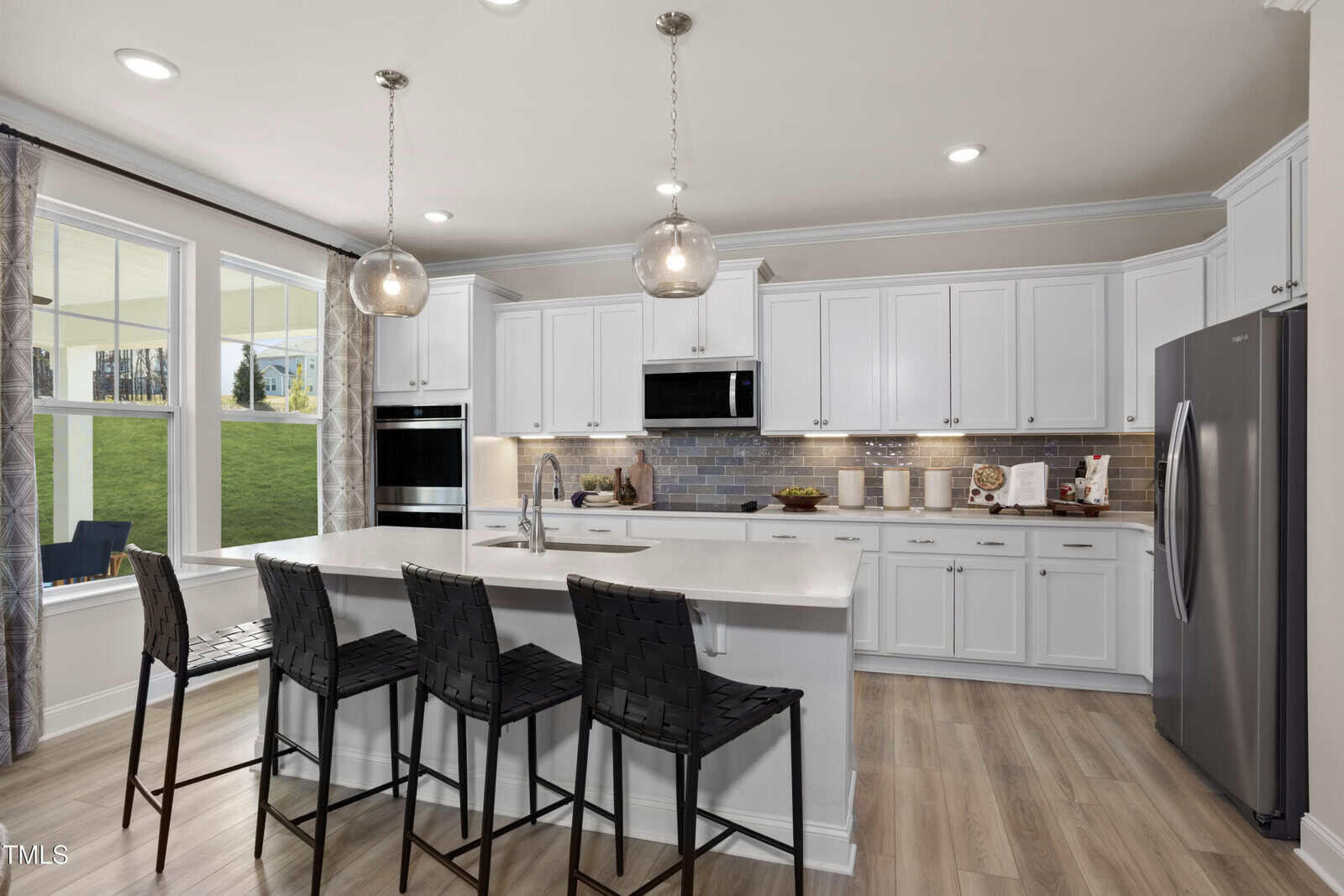 Modern kitchen with white cabinets, stainless steel appliances, a large island with sink, three black woven barstools, pendant lights, light wood flooring, and large windows overlooking a green lawn.