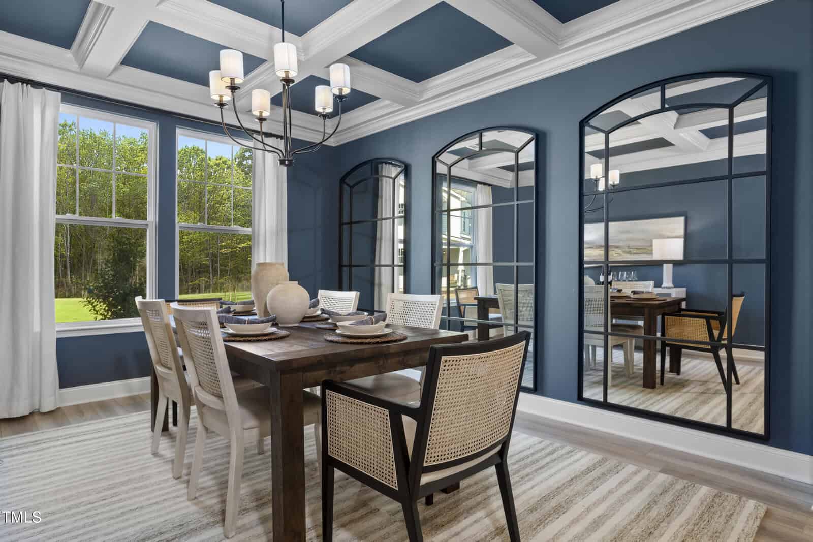 A modern dining room with a wooden table, six chairs, large arched mirrors on a blue wall, a chandelier, and tall windows with white curtains overlooking a green outdoor view.