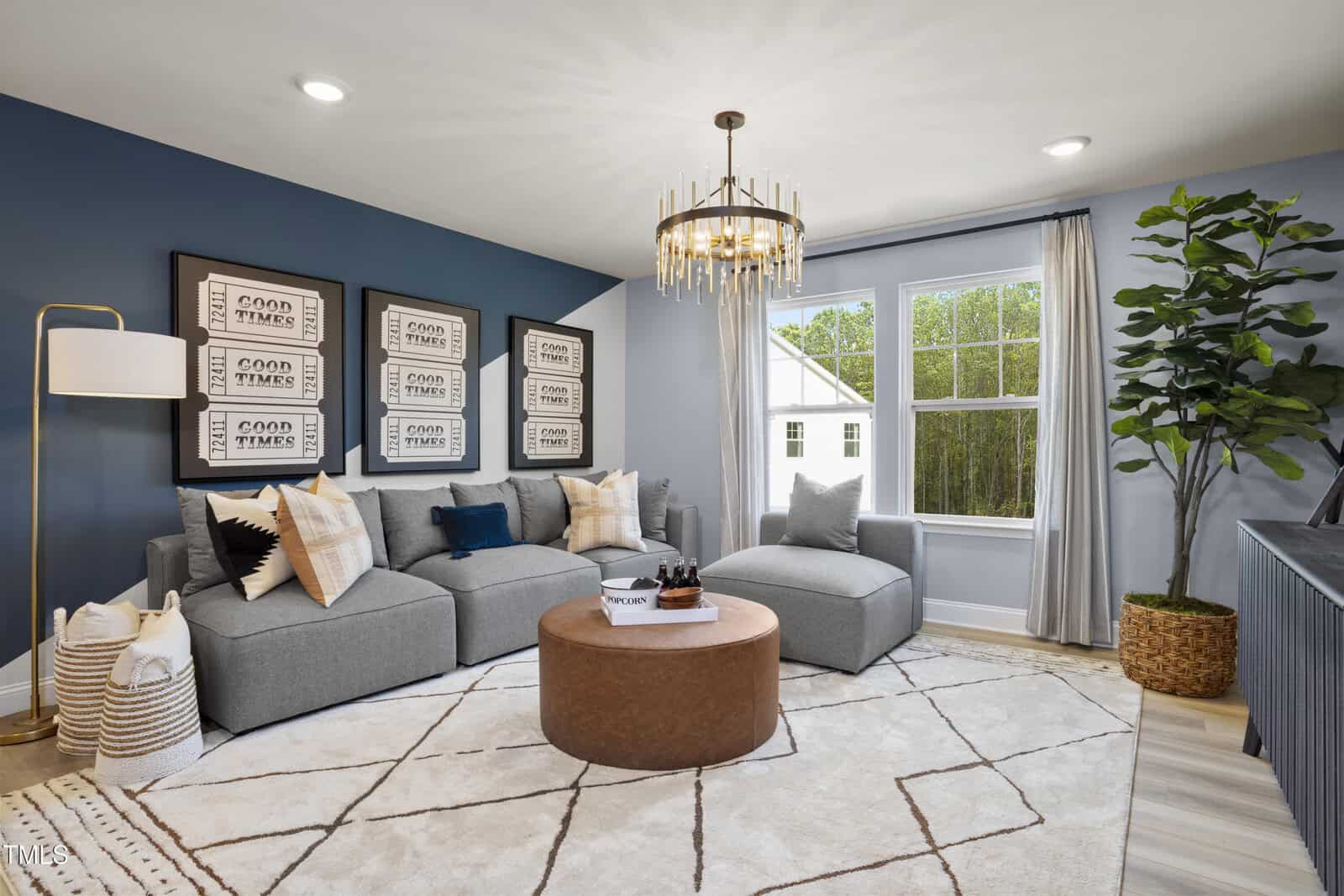 A modern living room with a grey sectional sofa, brown round ottoman, wall art, floor lamp, potted plant, large window with sheer curtains, and a geometric rug on light wood flooring.