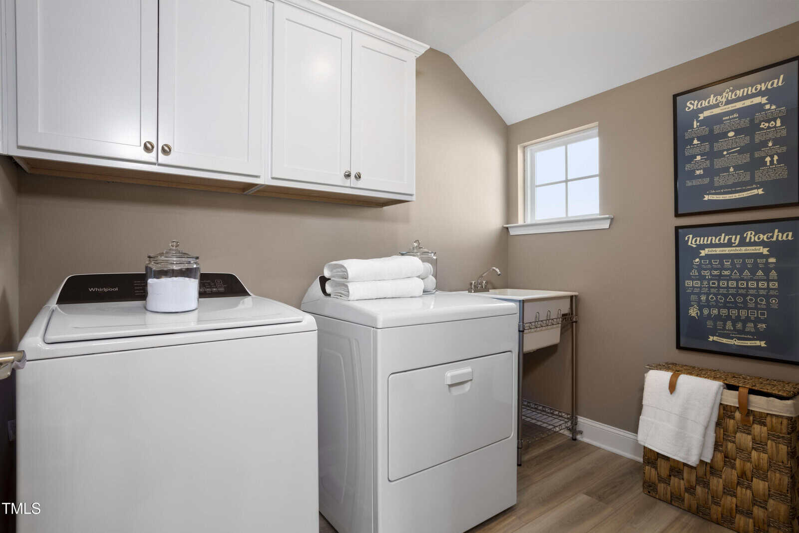 A modern laundry room with a washing machine and dryer, white cabinets above, a utility sink, a window, laundry-related wall art, a wicker basket, and neatly folded towels on the dryer.