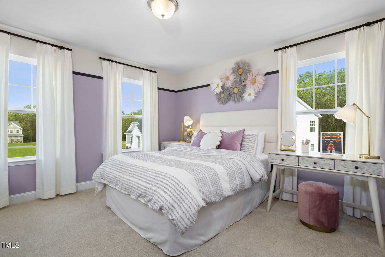 A cozy bedroom with light purple walls, a bed with white and gray bedding, a decorative flower arrangement above the headboard, two windows with white curtains, a small desk with a lamp, and a pink stool.