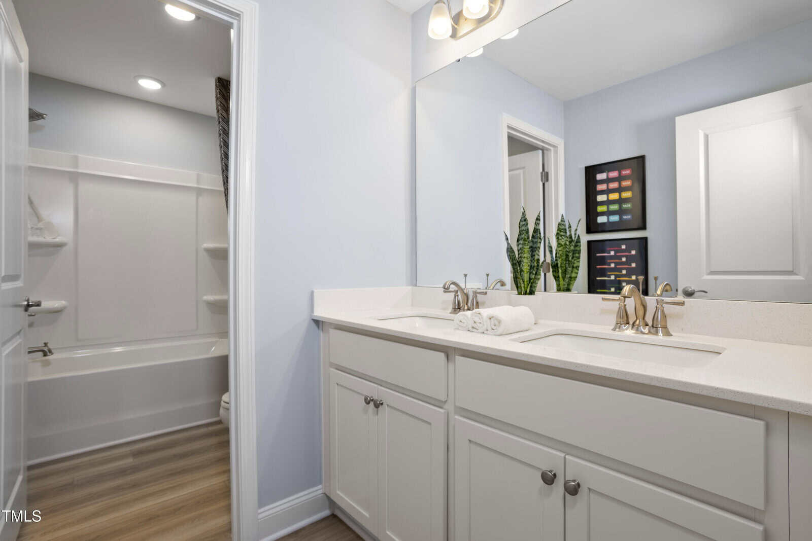 Modern bathroom with double sinks, white cabinets, large mirror, and gold fixtures. A plant, rolled towels, and colorful wall art decorate the space. In the background, there is a bathtub with built-in shelves.