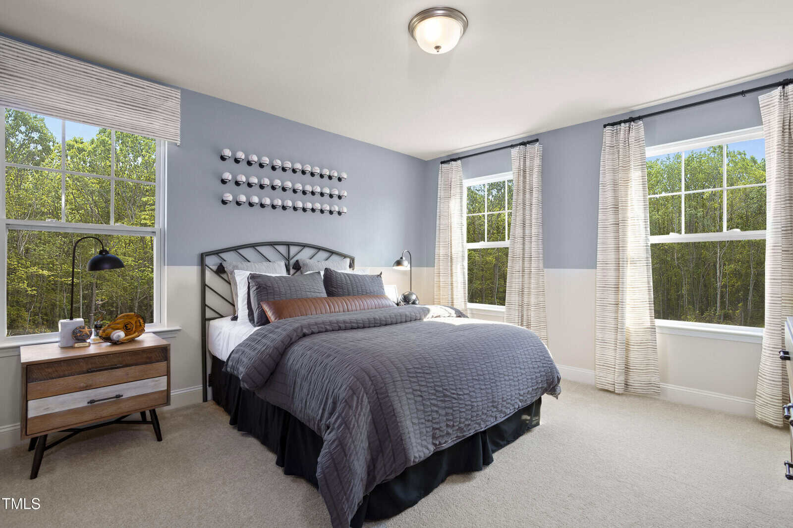 Modern bedroom with a gray and white color scheme, large bed with gray bedding, two windows with curtains, a nightstand with a lamp, and decorative wall art above the headboard. Sunlight streams in through the windows.