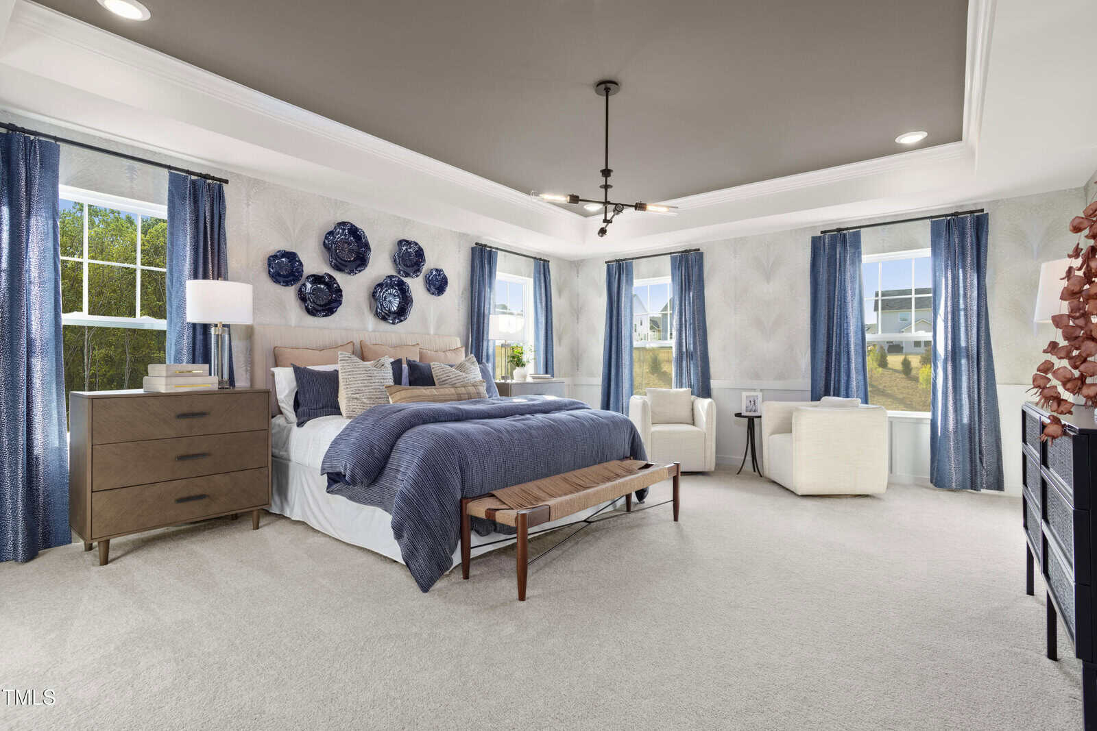 Bright, spacious bedroom with large windows, a bed with blue and beige bedding, wooden furniture, blue curtains, and navy wall art above the headboard. Two white chairs sit by the windows.