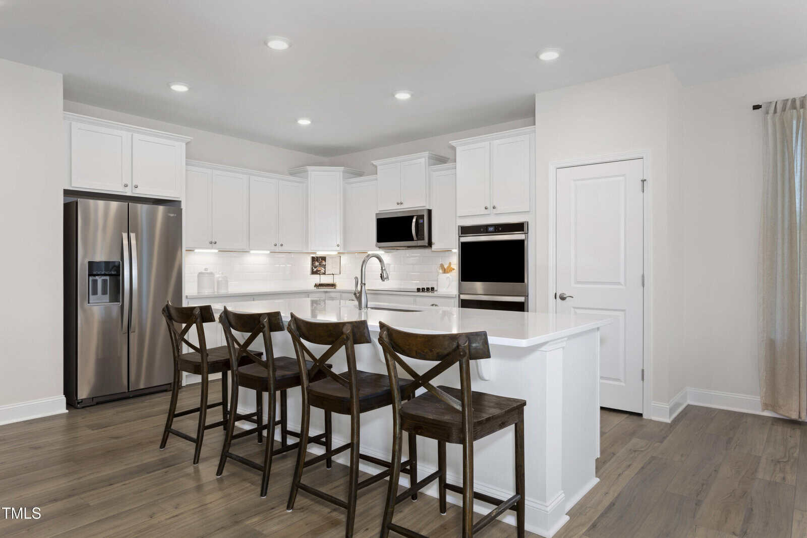 Modern kitchen with white cabinets, stainless steel appliances, an island with sink, and four wooden chairs. Recessed ceiling lights and light wood flooring. A window with beige curtains is visible on the right.