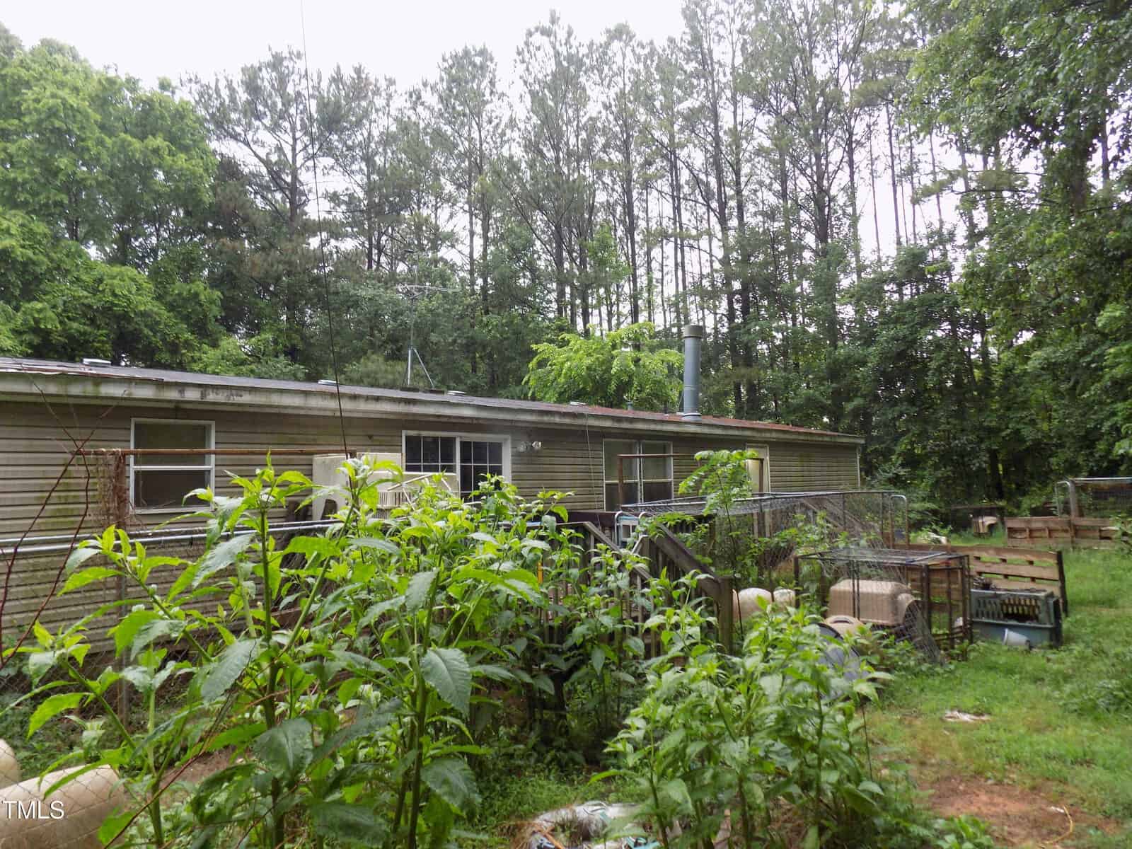 A weathered, single-story mobile home is surrounded by overgrown plants and trees. The yard has various items, including wire cages, wooden pallets, and garden vegetation, creating a cluttered, rustic appearance.