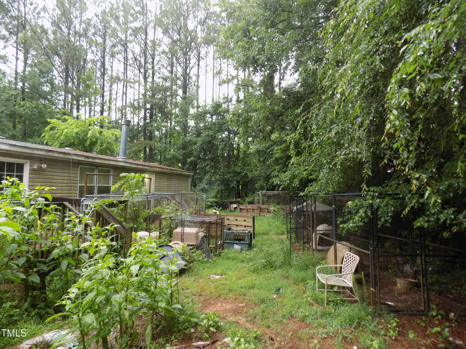 A fenced backyard with overgrown grass, scattered chairs, pet crates, and a tan house at the left. Trees and plants surround the area, and wire fencing sections off parts of the yard.