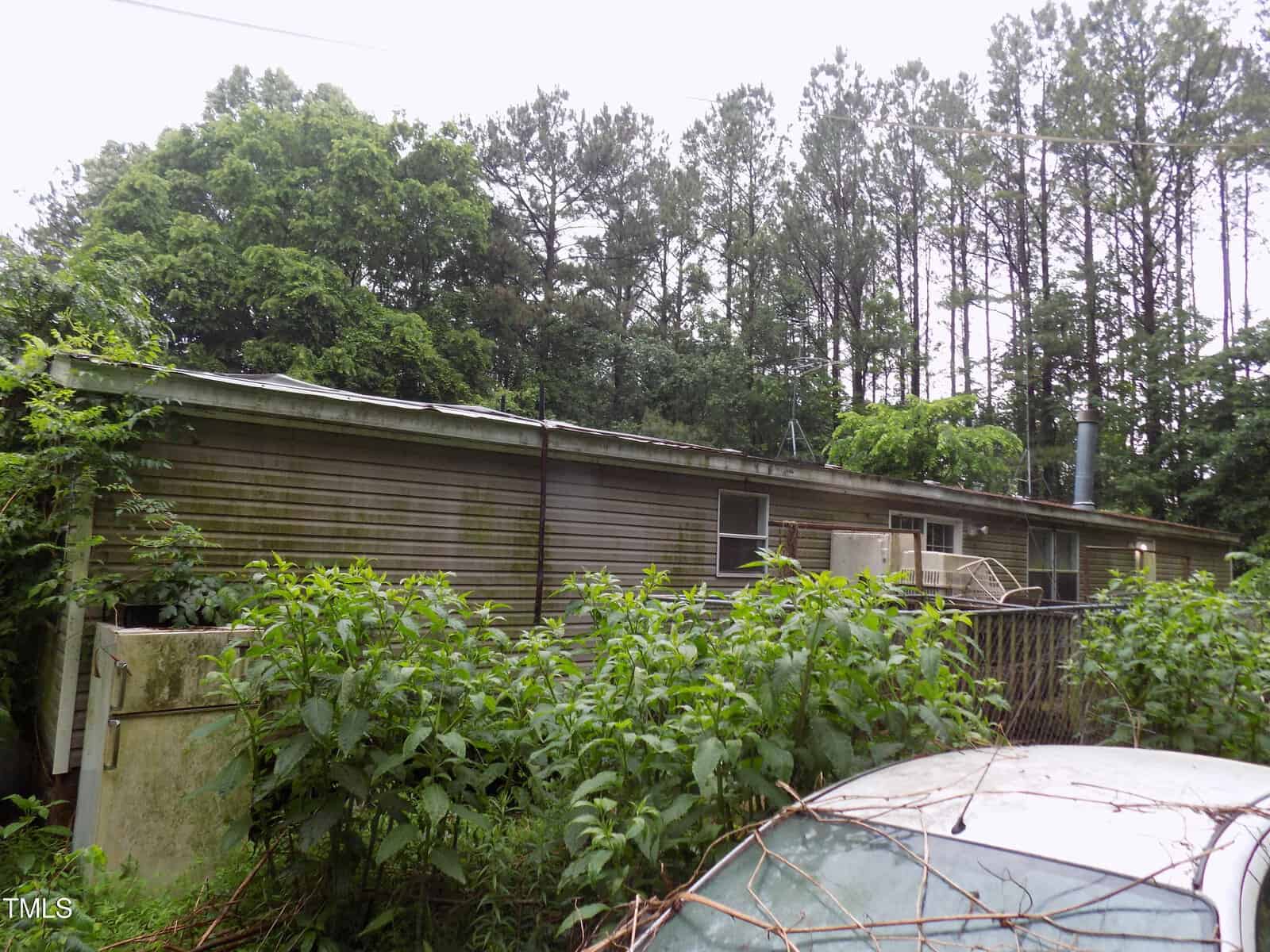 A single-story mobile home with weathered siding is surrounded by overgrown bushes and trees. An old air conditioning unit and other items are visible near the exterior. Tall pine trees are in the background.