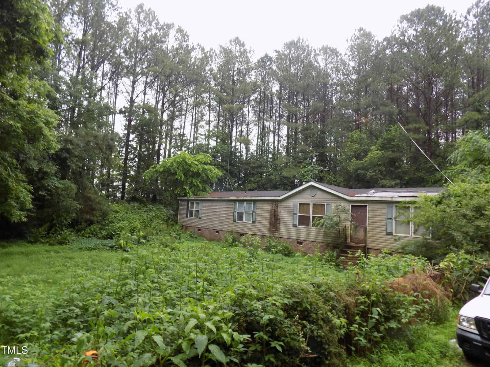 A weathered single-story mobile home sits amid overgrown grass and dense greenery, surrounded by tall pine trees under an overcast sky. Part of a white vehicle is visible on the right.