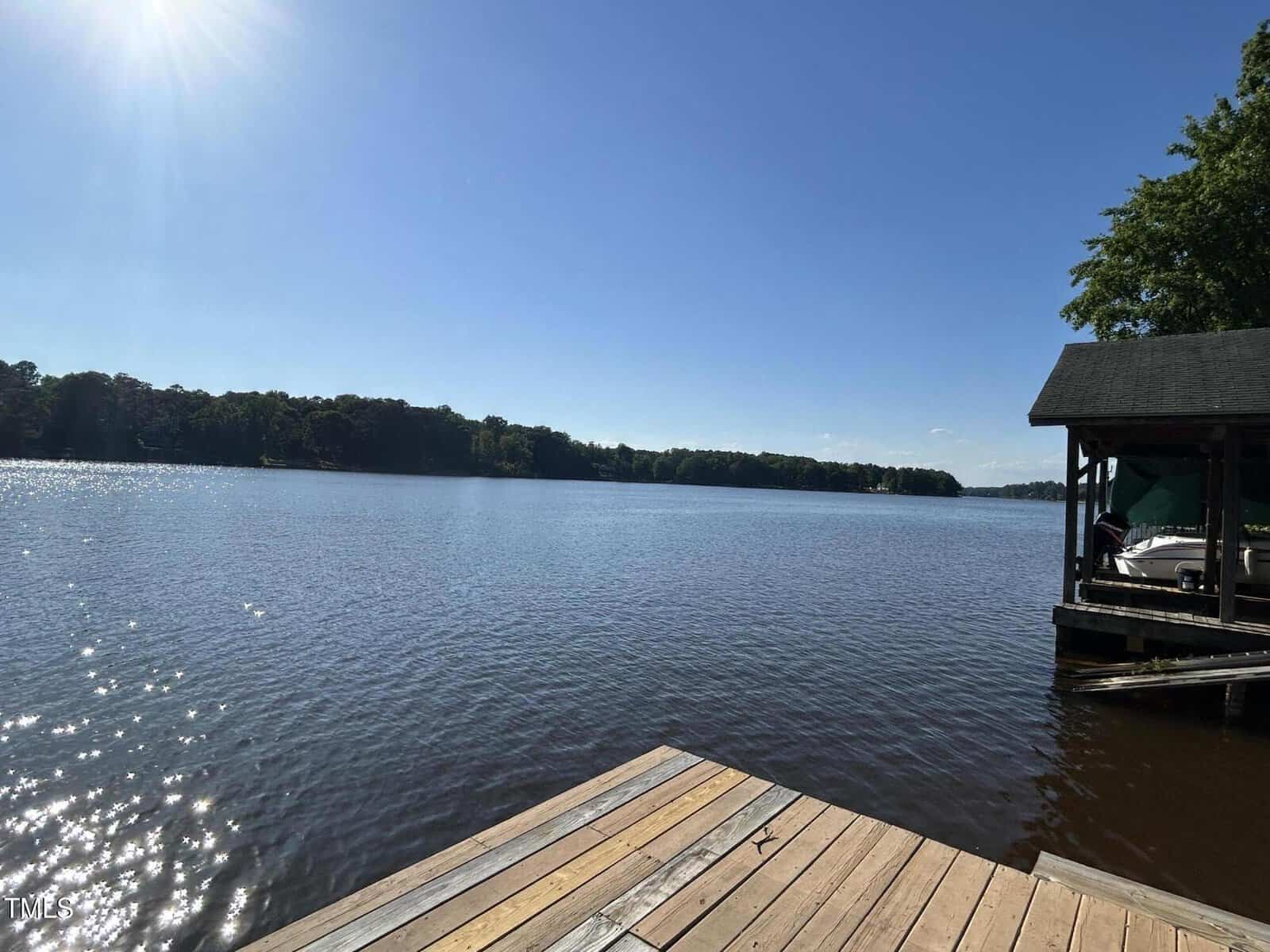 Sunny view of a calm lake with a wooden dock in the foreground and a covered boat slip to the right. Trees line the distant shoreline under a clear blue sky.
