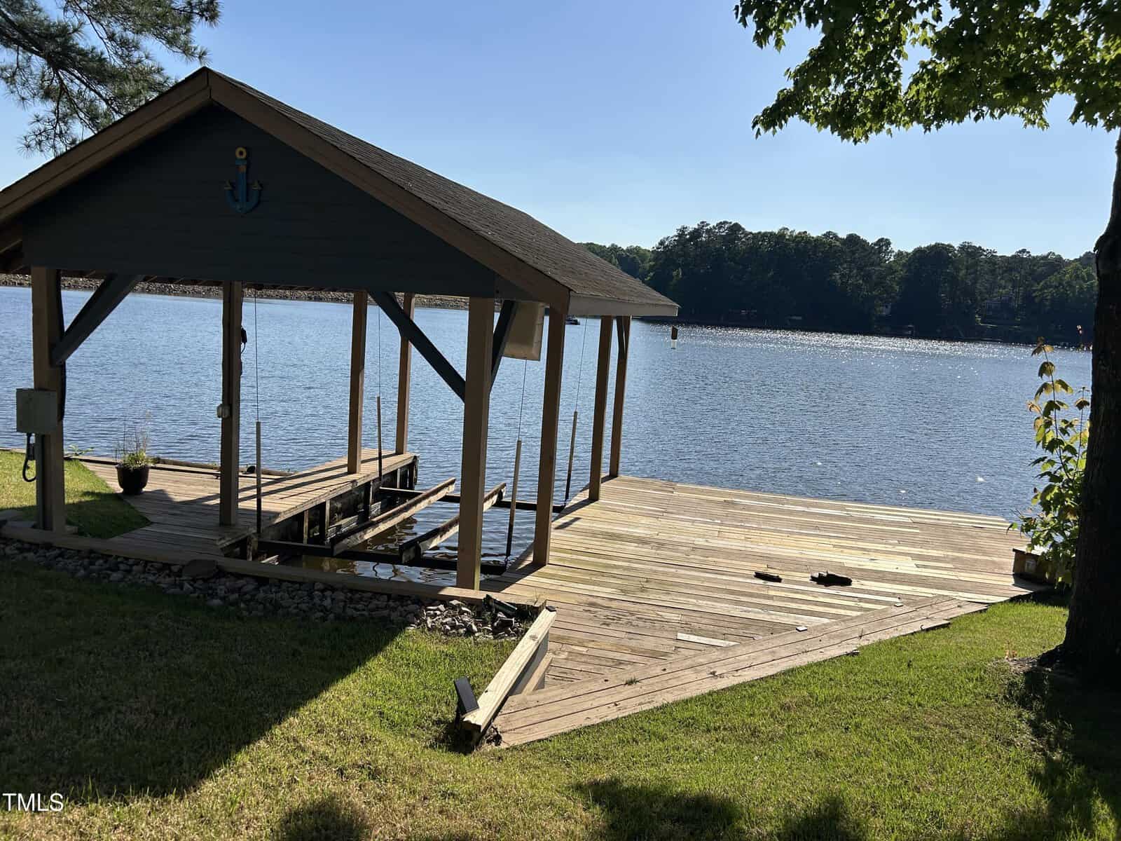 A wooden dock with a covered boat slip extends over a calm lake, surrounded by trees and greenery under a clear blue sky. Shadows from the trees and roof fall across the dock and grass.