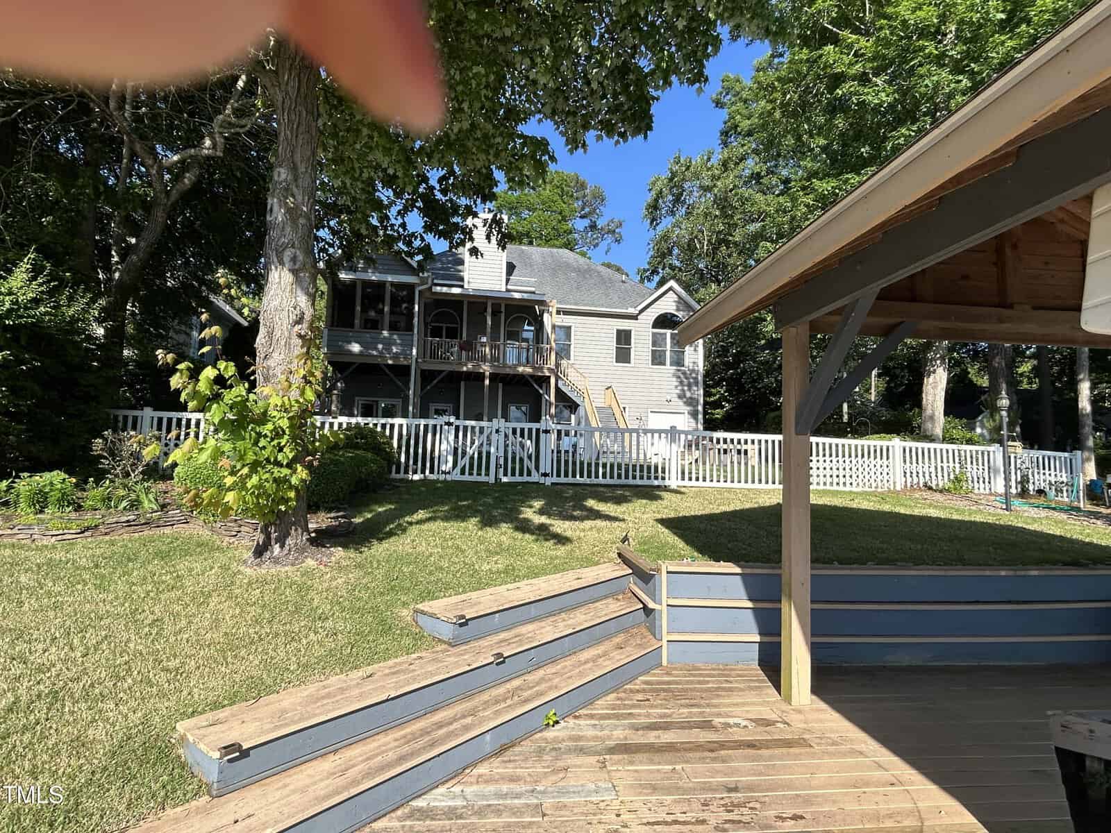 A large gray house with multiple balconies and a white picket fence sits in a spacious yard with green grass and trees. Wooden steps and part of a gazebo are visible in the foreground under a clear blue sky.