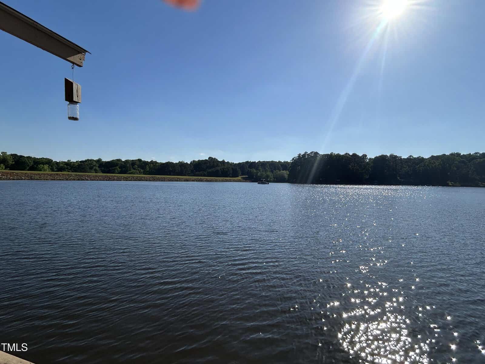 A lake with calm water under a clear blue sky. The sun is shining brightly in the upper right corner, and trees line the distant shore. Part of a rooftop and a hanging item are visible at the upper left.