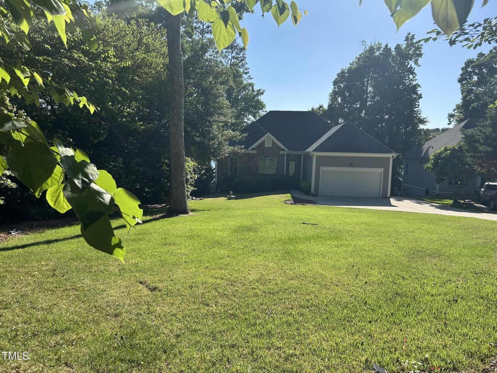 A suburban house with a dark roof, brick and siding exterior, and a two-car garage sits behind a large, sunny green lawn, surrounded by trees and neighboring homes. Leaves frame the top of the image.