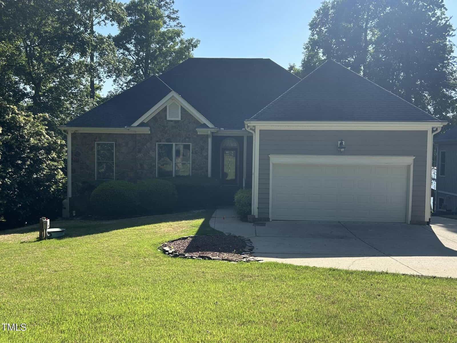 Single-story house with a two-car garage, stone and gray siding exterior, front porch, and well-kept green lawn. Trees and bushes surround the home, and the driveway curves gently to the garage.